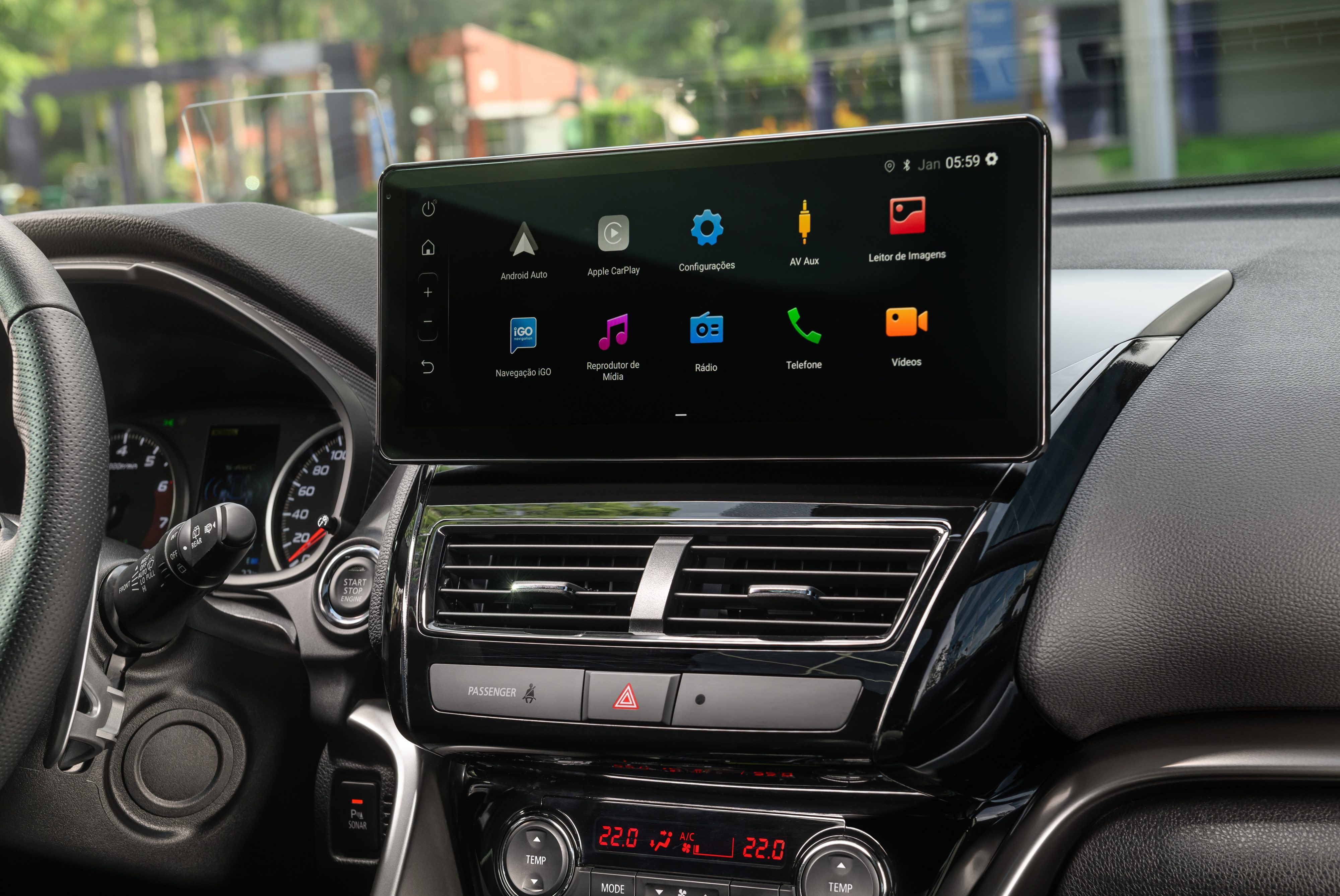 Painel interno de um carro com tela multimídia preta exibindo ícones de aplicativos como Android Auto, Apple CarPlay, Configurações, Rádio e Telefone. Abaixo, saídas de ar e controles de temperatura digitais marcando 22.0 graus. Volante e parte do painel de instrumentos são visíveis à esquerda