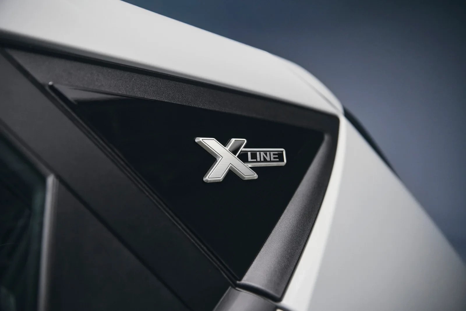 Emblema prateado X Line em destaque na lateral de um carro branco, sobre um fundo preto triangular e detalhes escuros