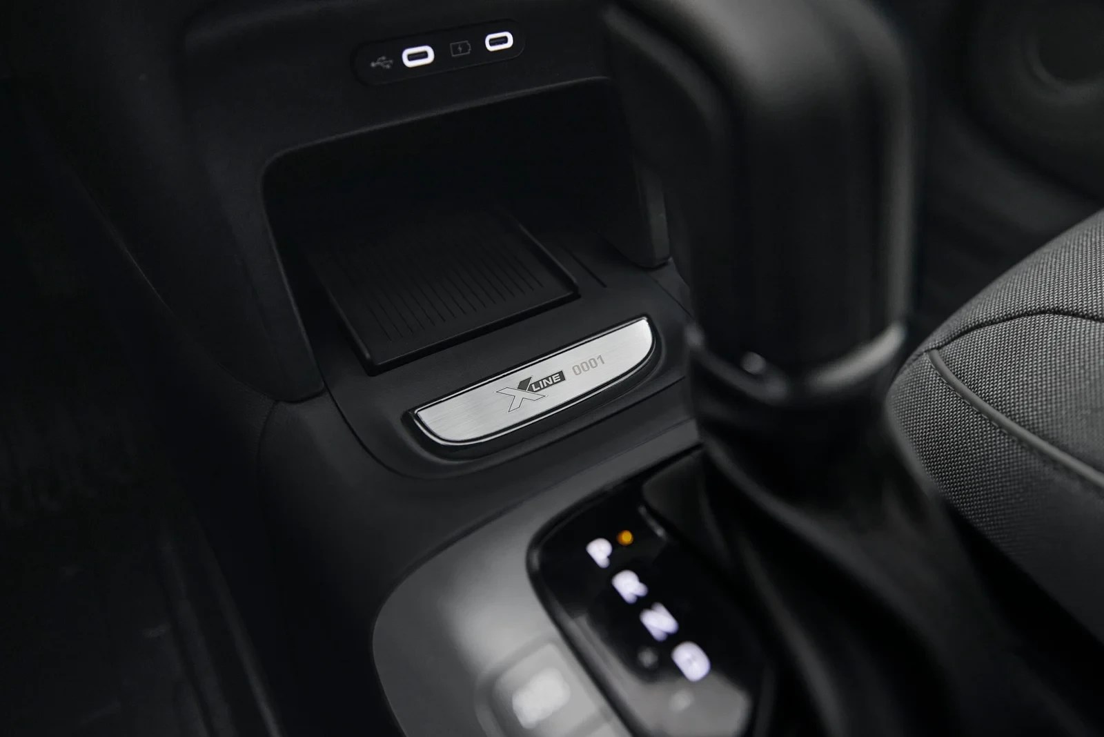 Interior de carro escuro, detalhe do console central com câmbio automático, porta-objetos e placa metálica X LINE 0001. Acima, duas portas USB-C e uma USB-A iluminadas. À direita, parte do assento do passageiro em tecido cinza escuro