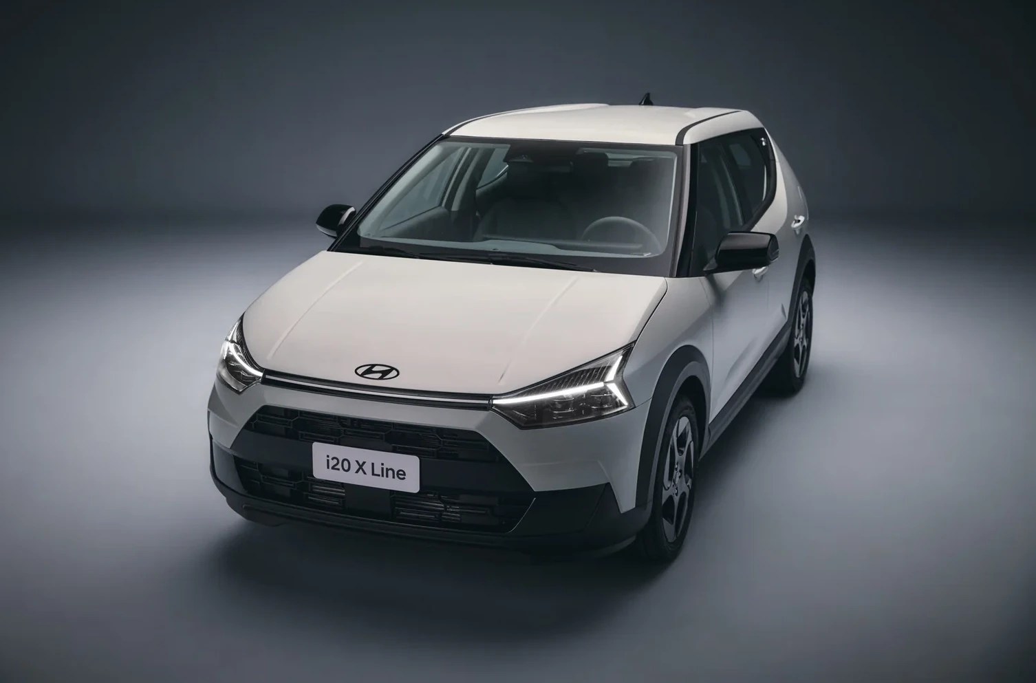 Carro Hyundai i20 X Line branco, visto de frente e ligeiramente de cima, com faróis acesos e detalhes pretos na grade e para-lamas, em um estúdio cinza