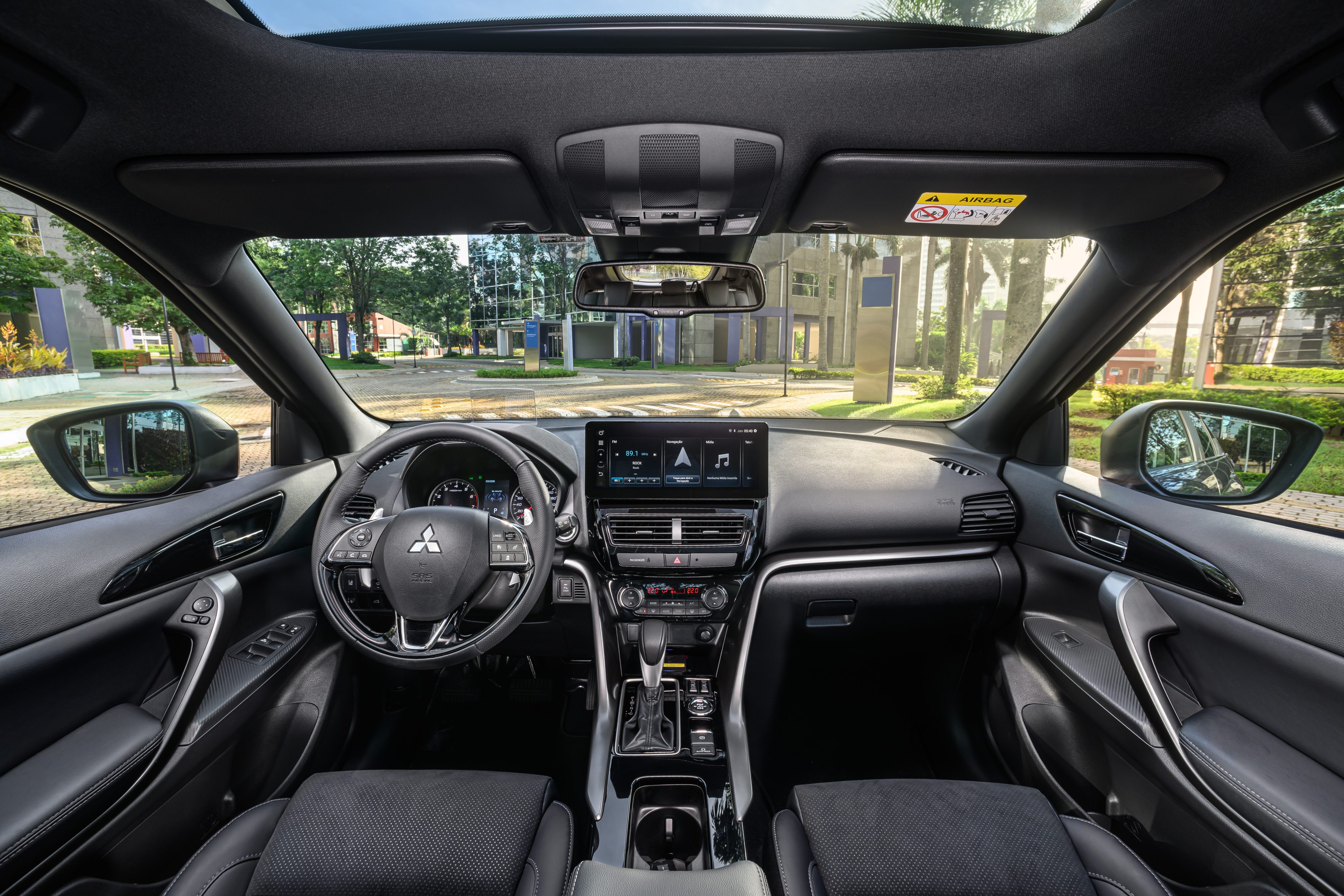 Interior de um carro moderno com bancos pretos, volante multifuncional e painel com tela multimídia. O para-brisa revela uma rua arborizada com prédios e postes sob luz solar