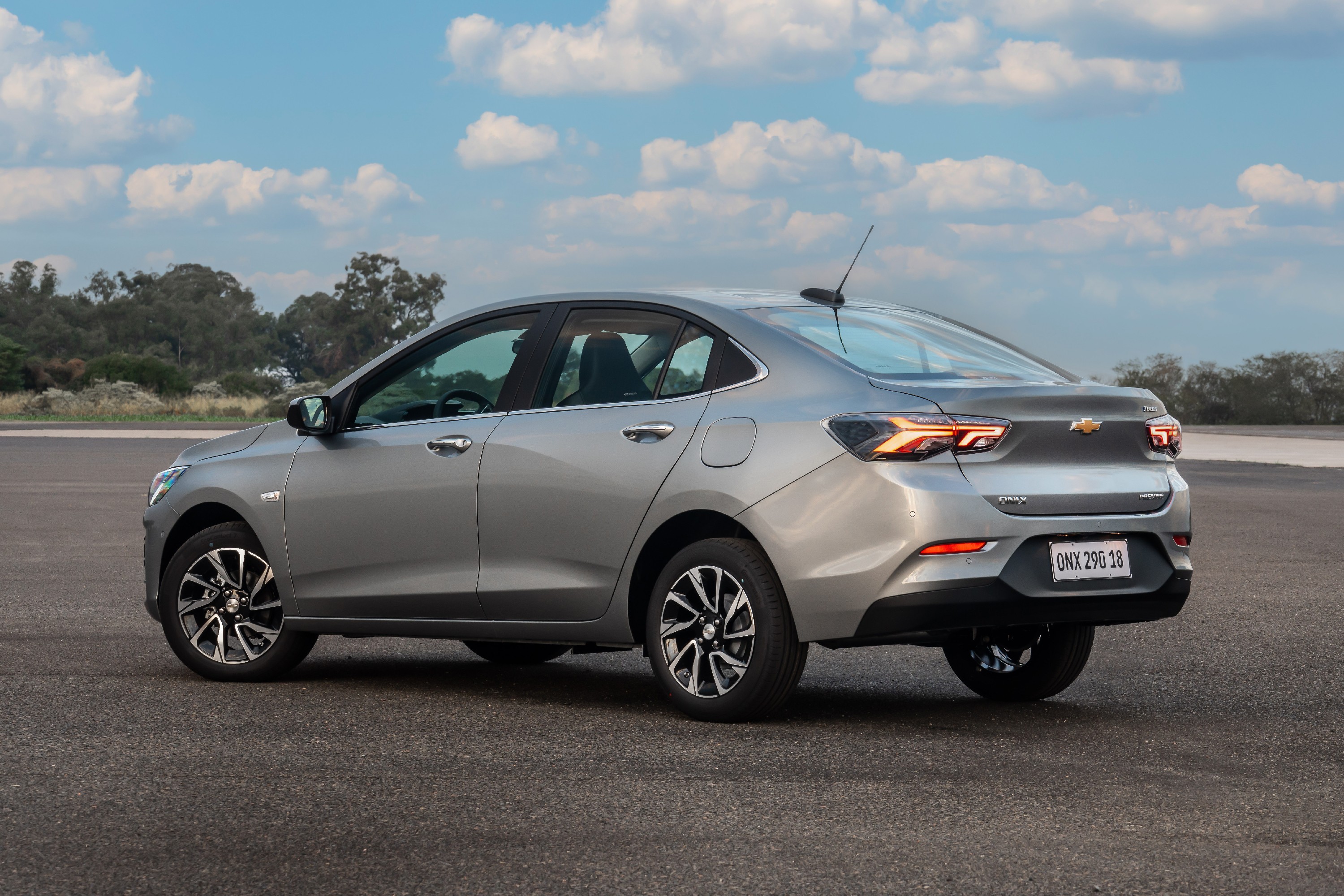 Carro Chevrolet Onix Plus sedan cinza claro, visto de trás e lateral, estacionado em asfalto com céu azul e nuvens ao fundo