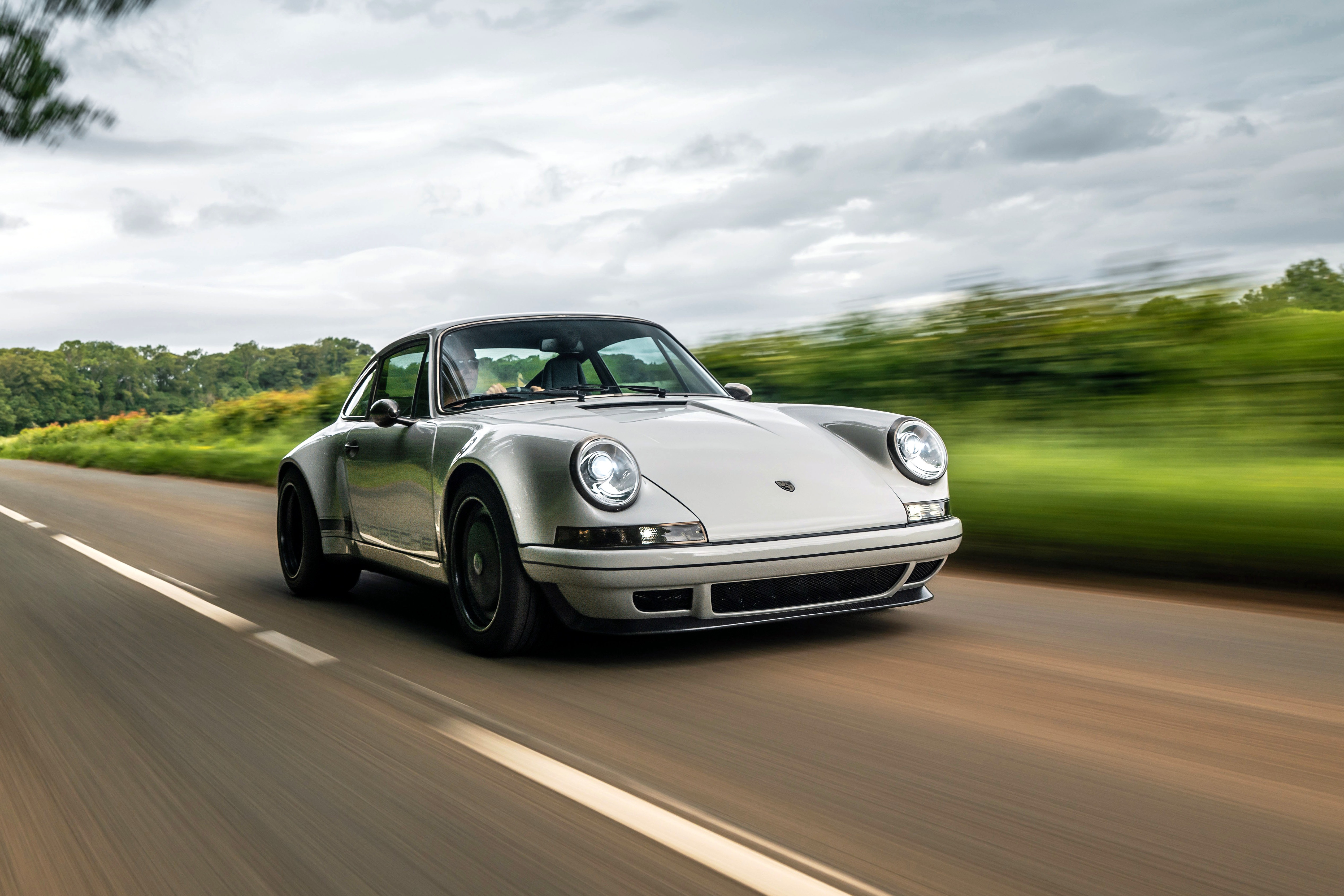 Carro esportivo prateado clássico, modelo Porsche 911, em movimento rápido numa estrada asfaltada. O fundo desfocado mostra vegetação verde e um céu nublado, transmitindo velocidade