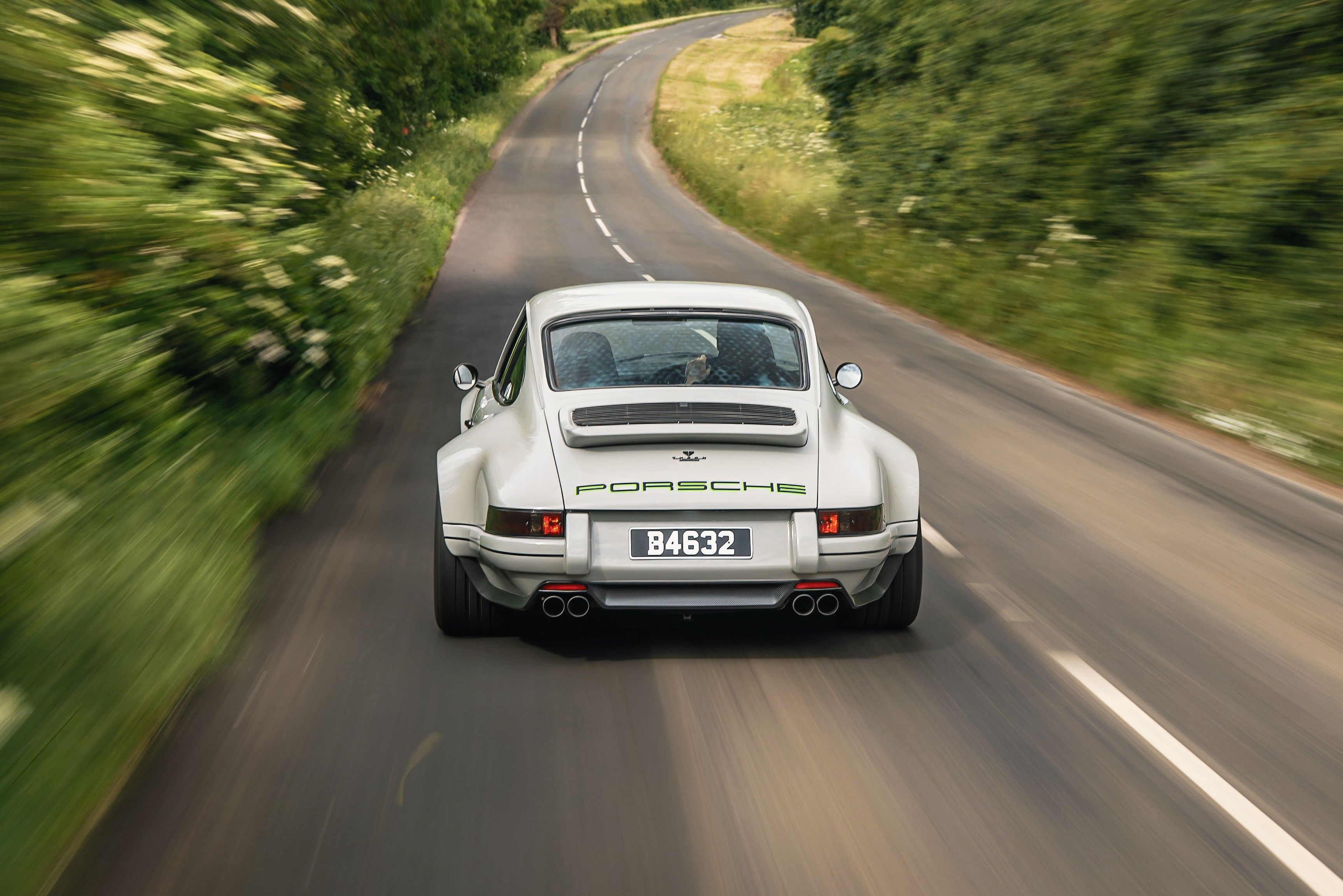 Porsche 911 branco clássico visto por trás, com a placa B4632, em uma estrada sinuosa cercada por vegetação verde borrada, indicando movimento
