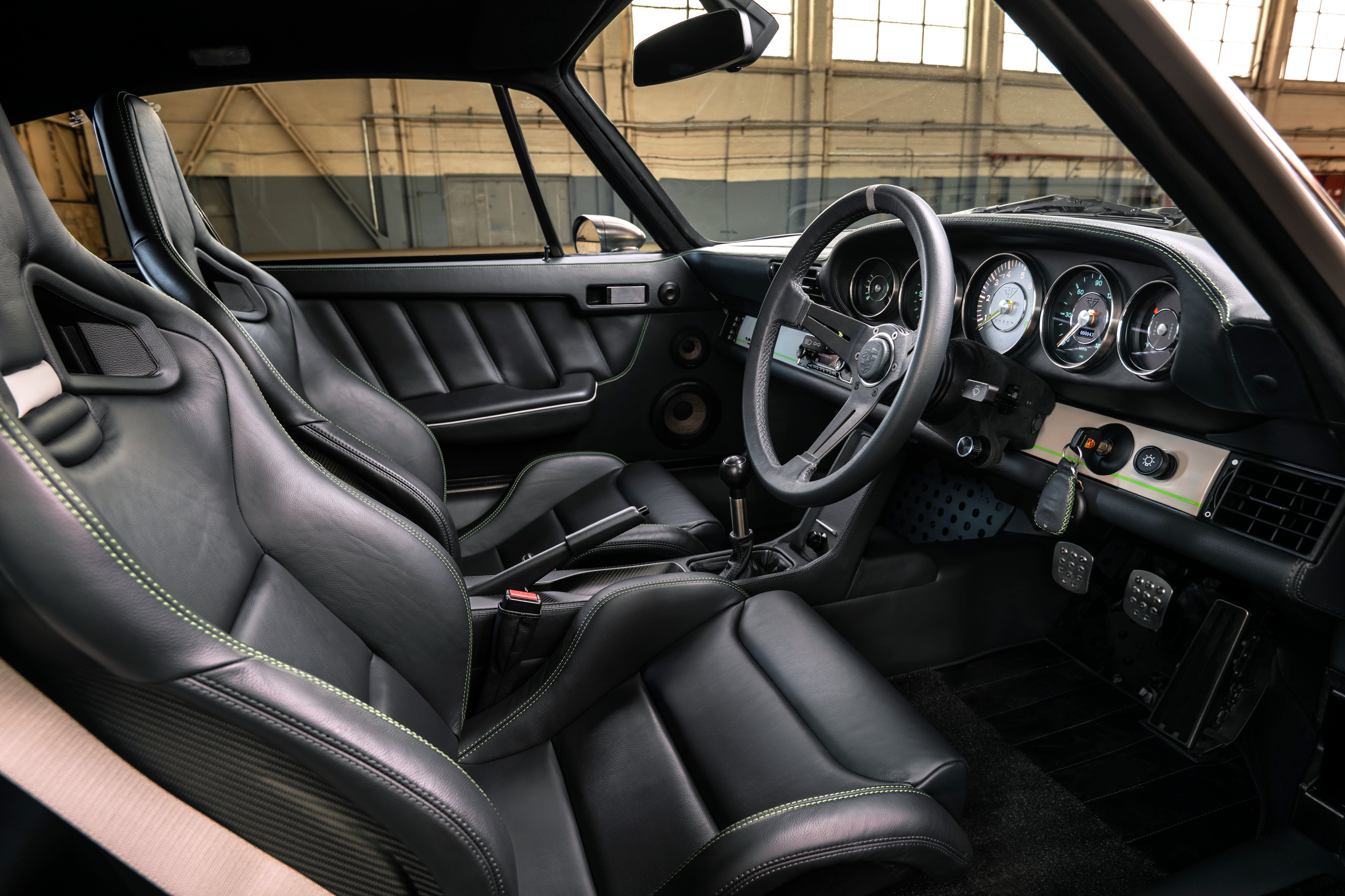 Interior de um carro esportivo com bancos de couro preto e costura verde, volante de três raios, painel com cinco mostradores redondos e pedais de metal perfurado. O carro está em um ambiente interno com janelas ao fundo