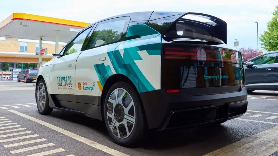 Carro el&eacute;trico branco e preto com adesivos Shell Recharge e Triple 10 Challenge na lateral, estacionado em frente a um posto de gasolina. O ve&iacute;culo tem um design futurista, com aerof&oacute;lio traseiro e rodas aerodin&acirc;micas. No para-choque traseiro, l&ecirc;-se Powering E-Motion