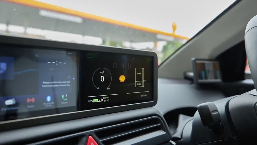 Painel digital de carro com velocímetro marcando zero, logo da Shell, indicador de bateria em 52% e autonomia de 44 milhas. Ao fundo, um posto de gasolina