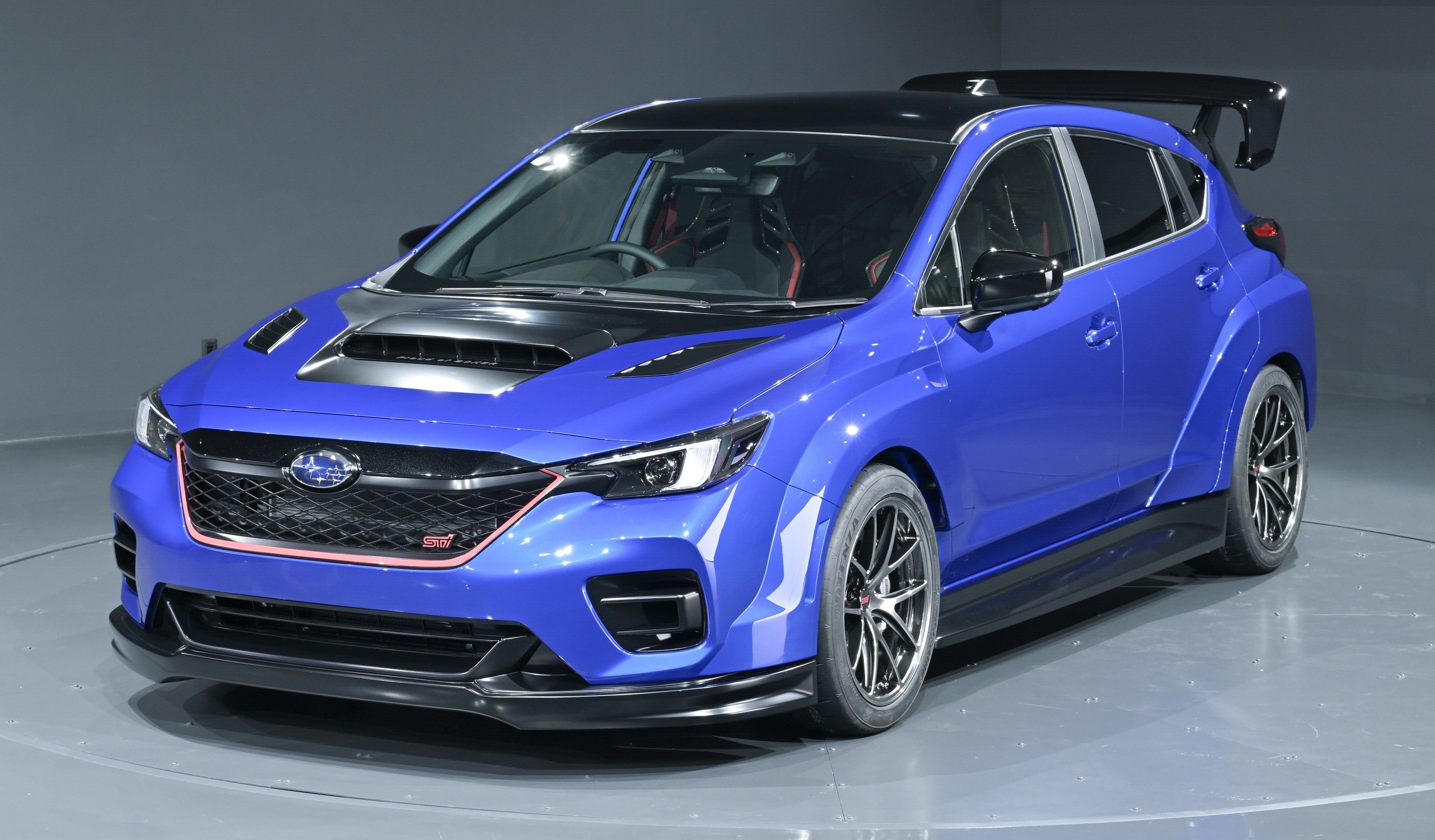 Carro esportivo azul metálico com capô preto e entradas de ar, grade frontal preta com contorno vermelho e emblema STI, rodas escuras e aerofólio traseiro, exibido em um estúdio com fundo cinza