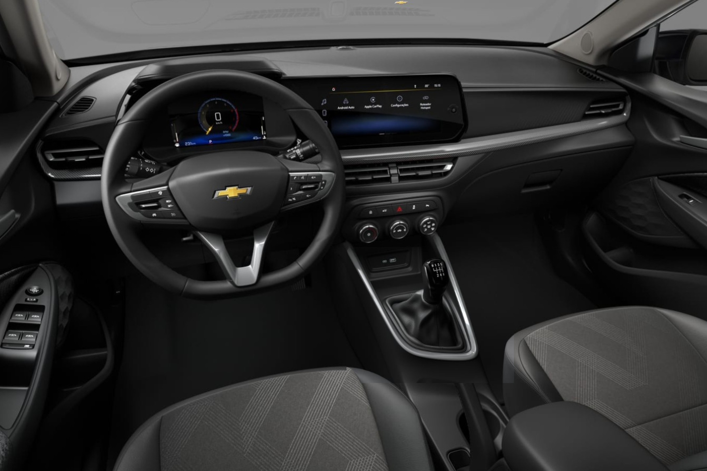 Interior de um carro moderno, com volante multifuncional preto e logo dourado da Chevrolet. Painel digital exibe velocímetro e tela multimídia sensível ao toque. Câmbio manual de cinco marchas e bancos de tecido cinza escuro com detalhes geométricos. O ambiente é predominantemente preto e cinza, com detalhes prateados no painel e console central