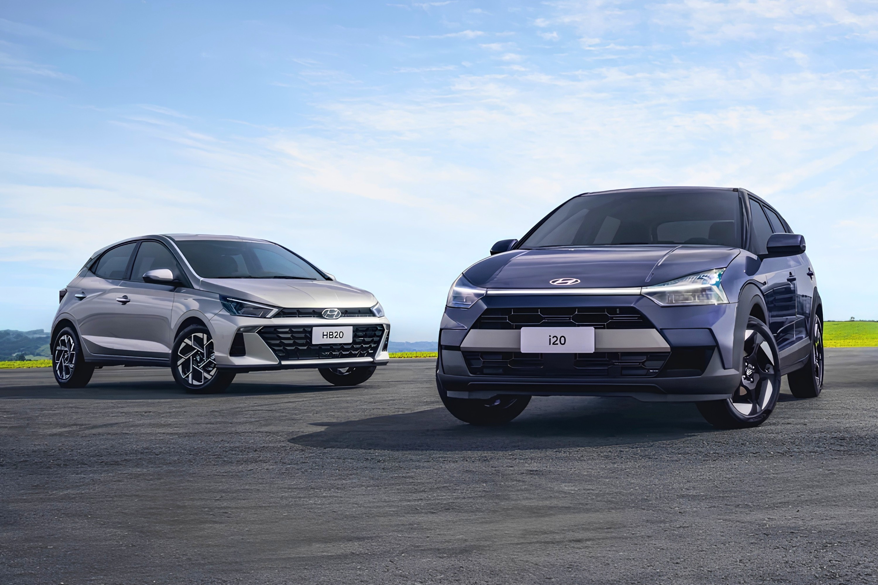 Dois carros Hyundai em uma pista de asfalto sob céu azul. À esquerda, um HB20 prata, e à direita, um i20 azul escuro com detalhes pretos, ambos vistos de frente