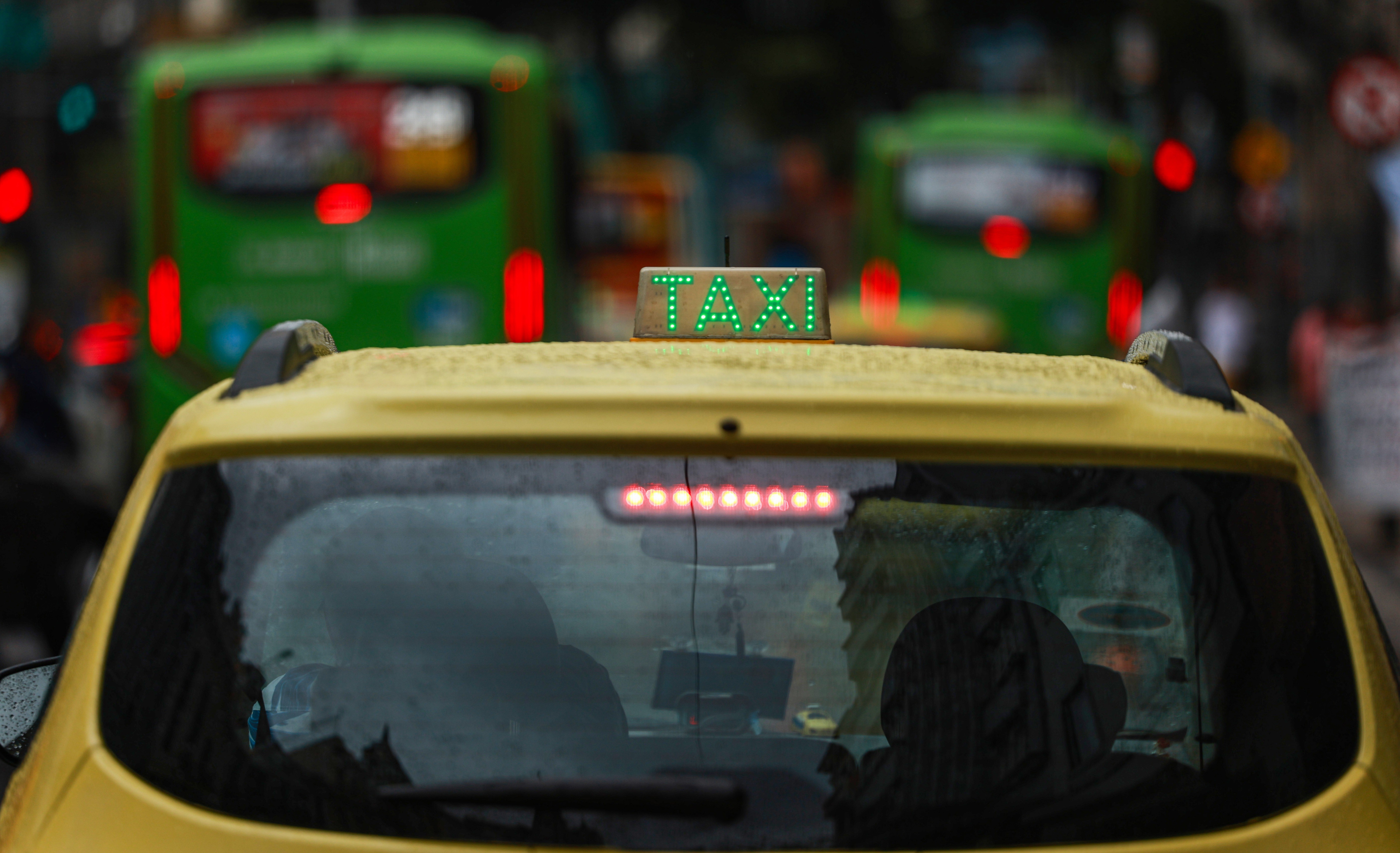 Traseira de um táxi amarelo com o letreiro TAXI em verde luminoso no teto, em meio ao trânsito urbano. Gotas de chuva cobrem o vidro traseiro e o teto do carro. Ao fundo, ônibus verdes e luzes vermelhas de freio estão desfocados, indicando movimento em uma rua movimentada
