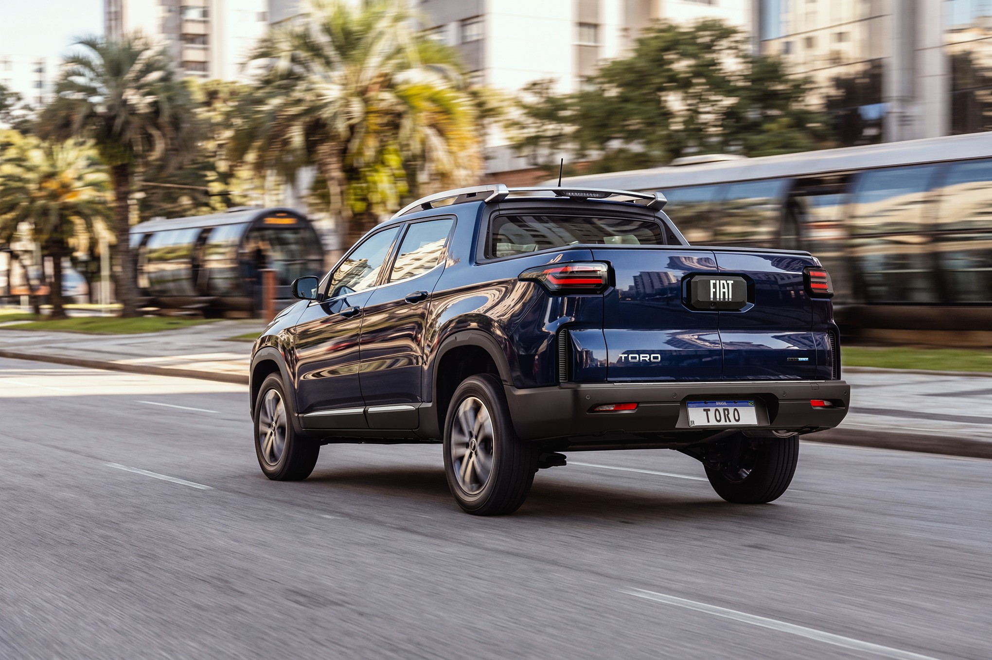 Picape Fiat Toro azul escura em movimento, vista de traseira e lateral, em uma rua urbana com palmeiras e pr&eacute;dios ao fundo, e um &ocirc;nibus borrado passando