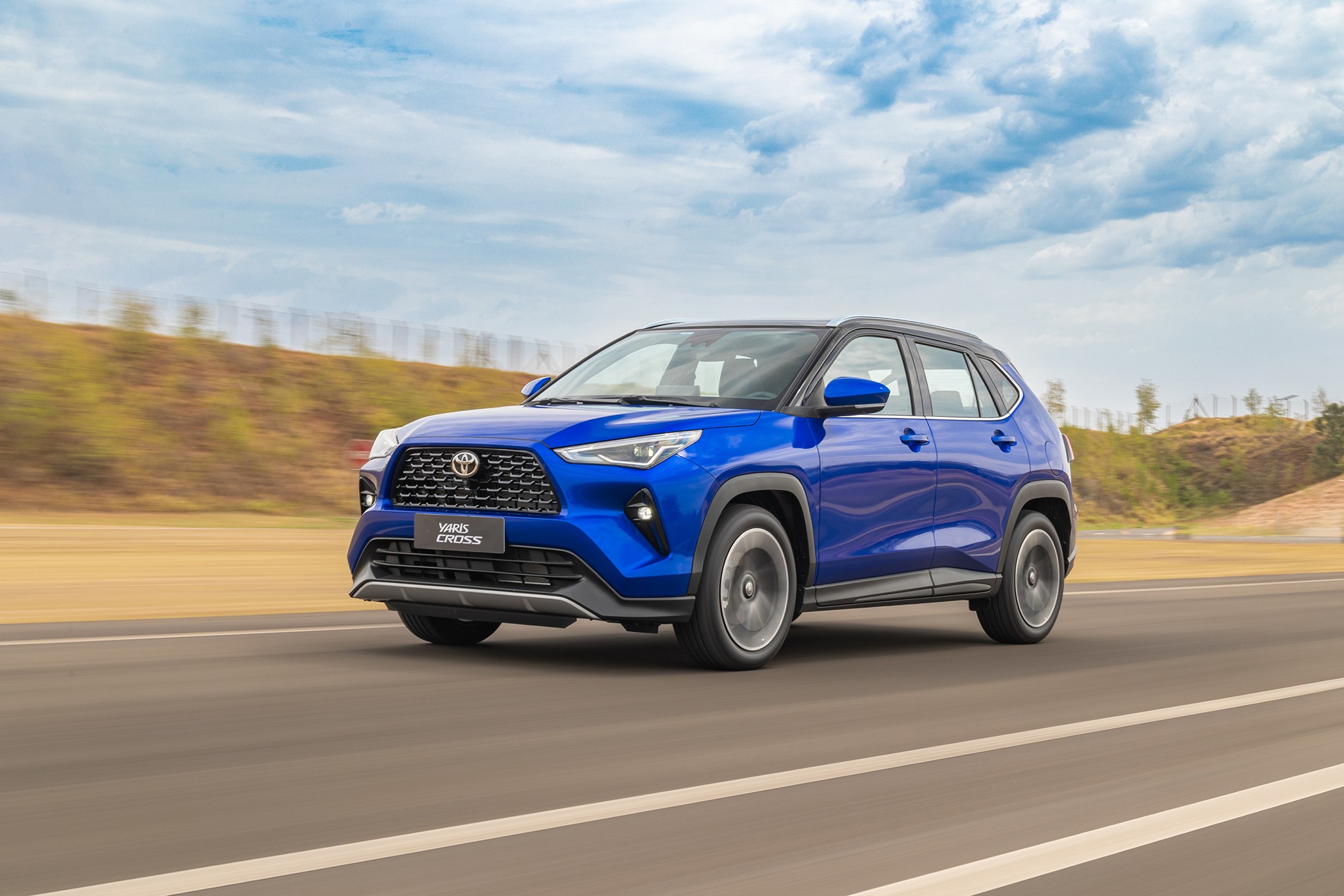 SUV Toyota Yaris Cross azul vibrante em movimento, visto de frente e lateral, em uma estrada asfaltada com faixas brancas. O céu tem nuvens esparsas e o fundo mostra colinas com vegetação.