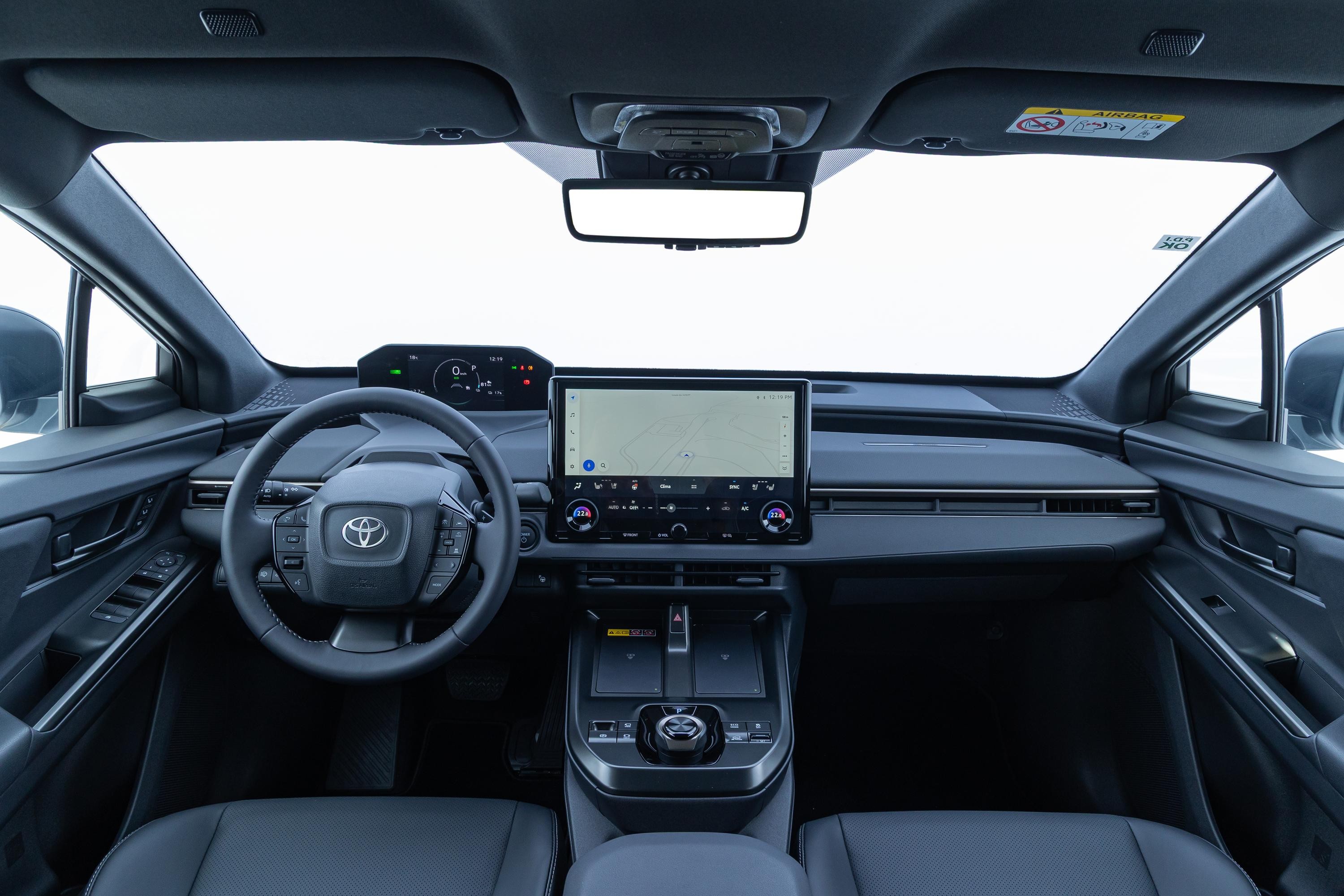 Interior de um carro moderno, com painel escuro e volante multifuncional com o logo da Toyota. Há uma tela multimídia grande no centro e um painel de instrumentos digital acima do volante. O para-brisa e as janelas laterais mostram um fundo branco, sugerindo um estúdio ou céu claro. Os bancos são de couro escuro e o console central possui botões e porta-objetos