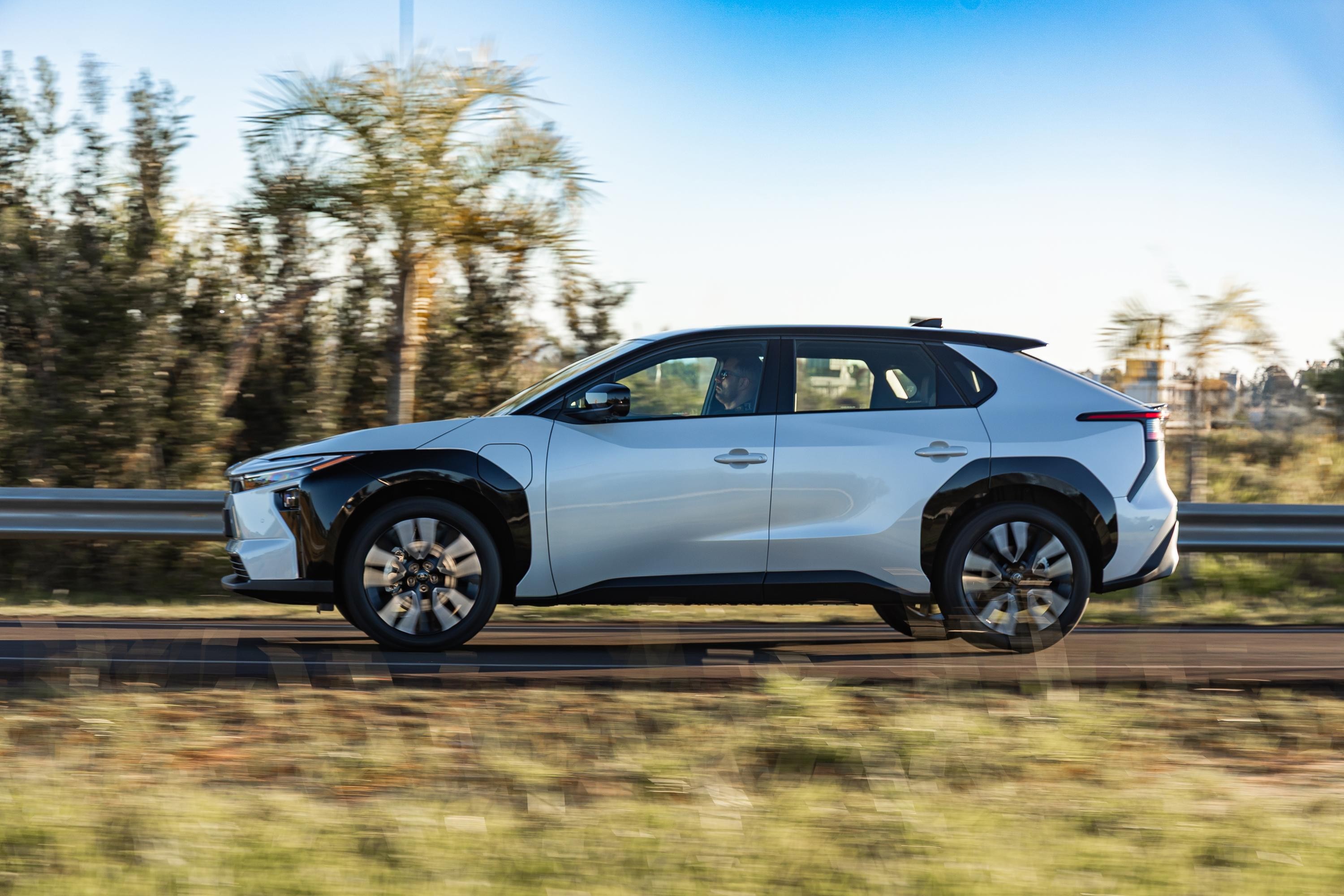 SUV elétrico branco e preto em movimento, visto de perfil, com fundo desfocado de árvores e céu azul