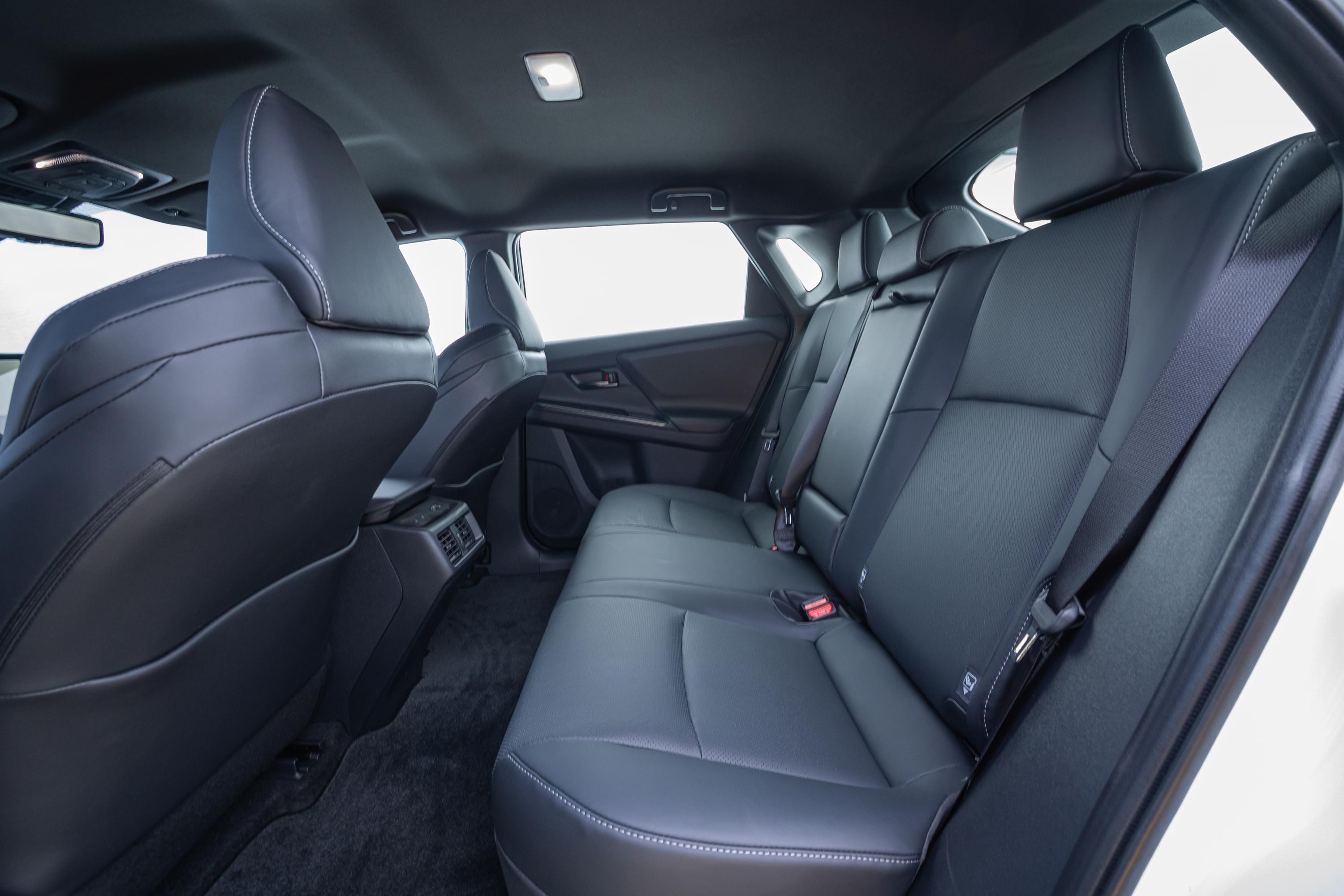 Interior de um carro com bancos traseiros de couro preto, costura branca e cintos de segurança vermelhos. O teto é escuro, com uma luz acesa. As janelas laterais mostram um fundo branco. O piso é forrado com carpete escuro.