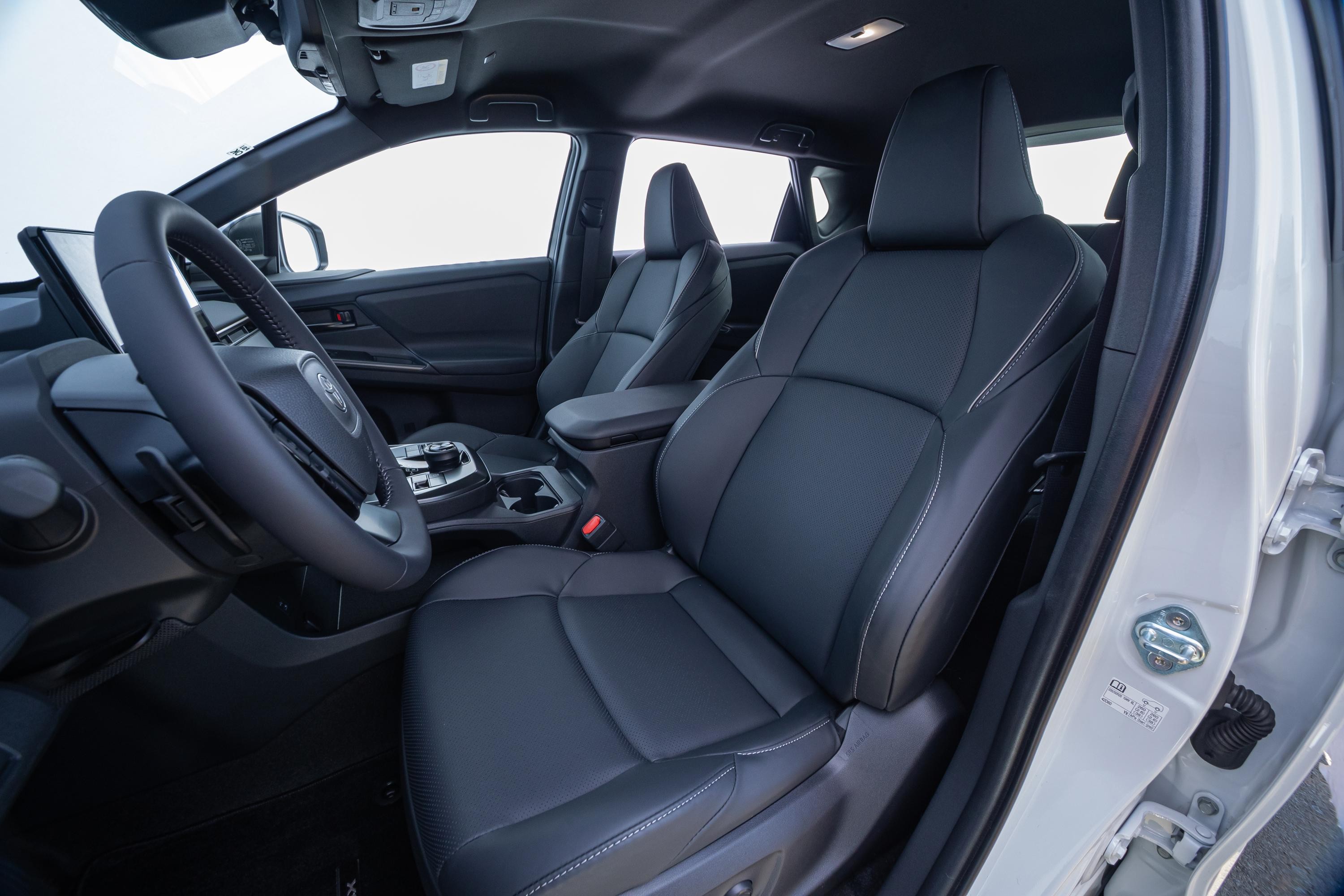 Interior de um carro moderno com bancos de couro escuro e costuras brancas, volante à esquerda, console central com porta-copos e tela digital no painel. A porta do passageiro está aberta, revelando a estrutura interna do veículo.
