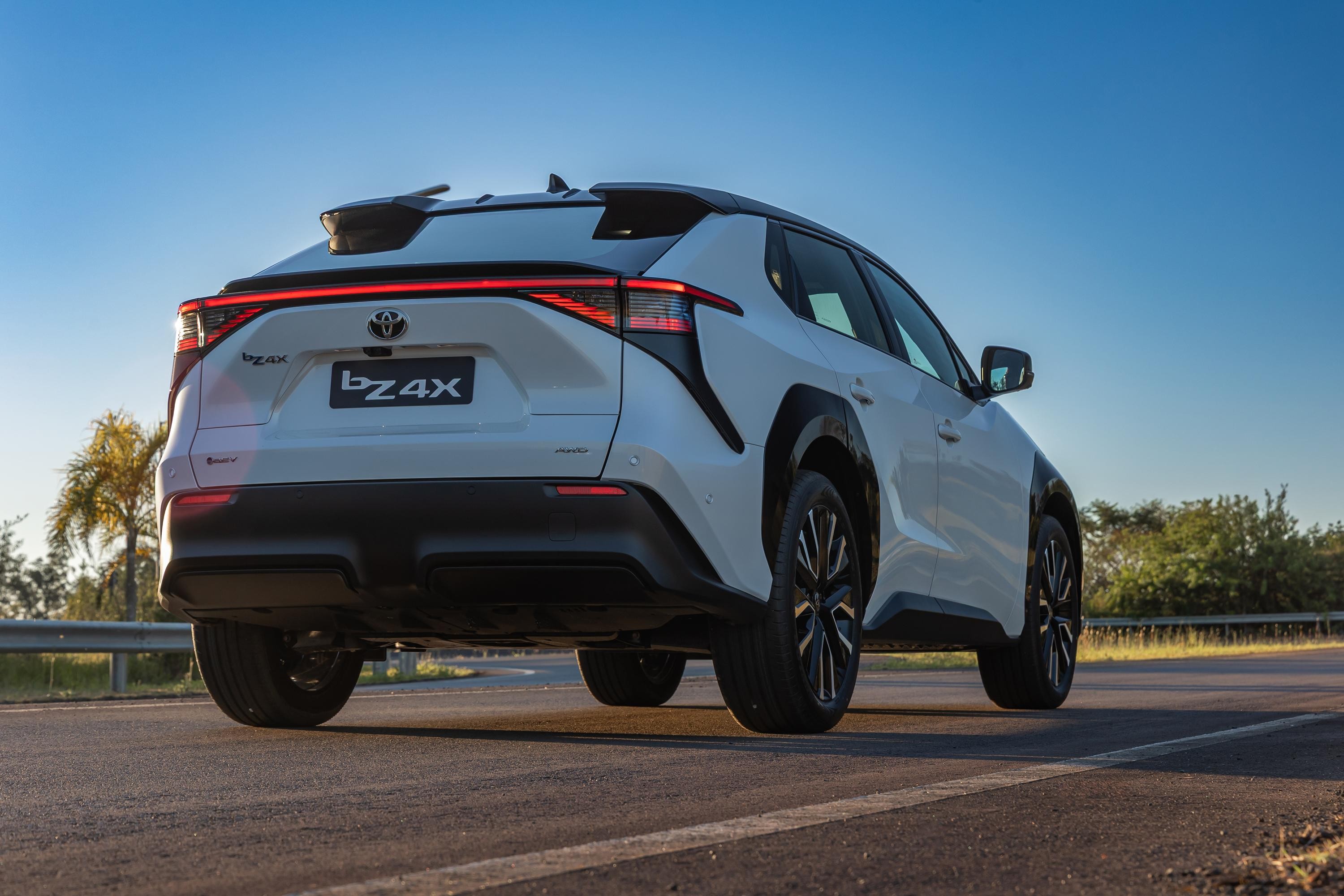 SUV Toyota bZ4X branco, visto por trás, parado em uma estrada asfaltada com faixa branca. O carro tem lanternas traseiras em LED vermelhas, para-choque preto e rodas escuras. Ao fundo, céu azul claro e vegetação verde com uma palmeira à esquerda