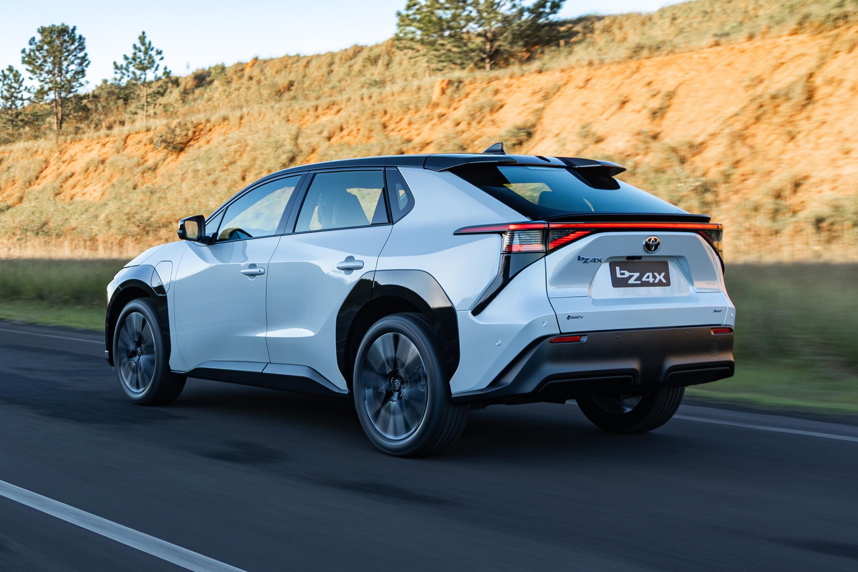 SUV elétrico Toyota bZ4X branco em movimento, visto de traseira lateral, com rodas escuras e teto preto, em estrada asfaltada com faixa branca. Ao fundo, um morro com vegetação seca e algumas árvores verdes no topo