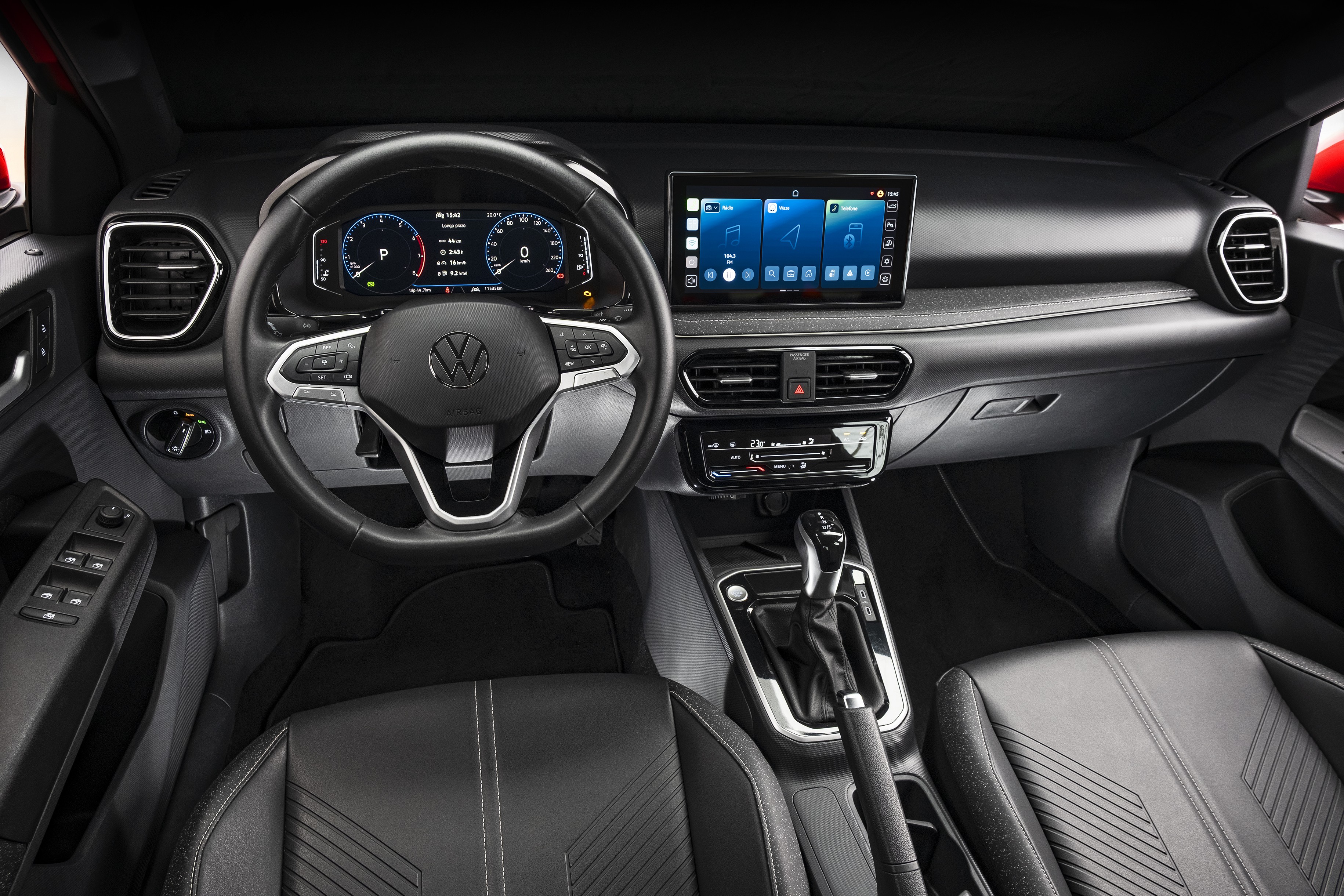 Interior de um carro moderno com painel escuro, volante multifuncional com logo Volkswagen, painel digital e tela multimídia central exibindo ícones azuis. O console tem câmbio automático, freio de mão e controles de ar condicionado. O banco do motorista é de couro preto com costuras brancas e detalhes geométricos