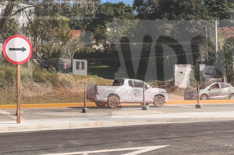 Sinal de trânsito circular branco e vermelho com seta preta apontando para a direita, em uma rua com asfalto novo e marcações brancas. Ao fundo, uma picape camuflada e um carro cinza estacionados, com vegetação e casas ao redor sob céu azul