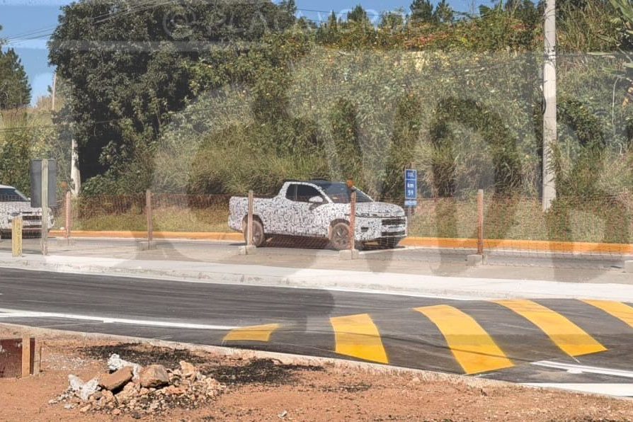 Duas picapes camufladas, uma branca e outra cinza, estacionadas em uma rua com faixa de pedestres amarela e branca, ladeada por vegetação densa e um poste de energia, sob céu azul claro