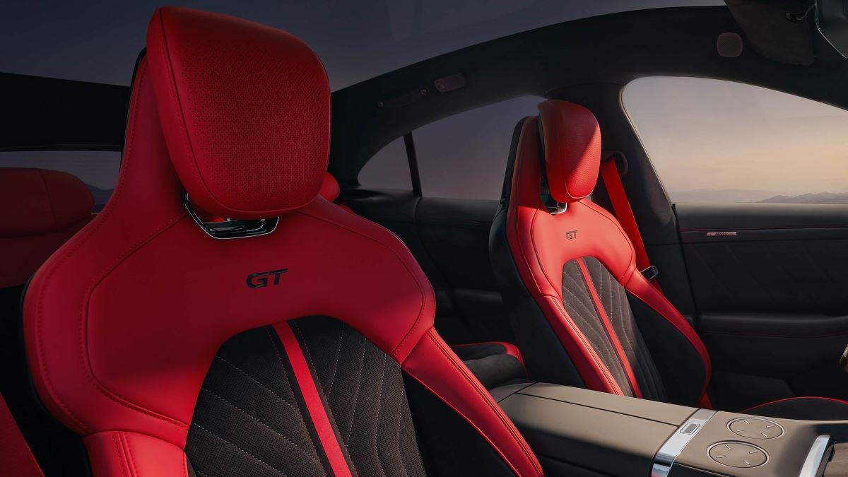 Interior de carro esportivo com bancos de couro vermelho e detalhes pretos, costura em losango e o logo GT bordado. O console central cinza claro com botões redondos e detalhes cromados. Ao fundo, uma paisagem montanhosa sob um céu claro ao entardecer, visível pela janela