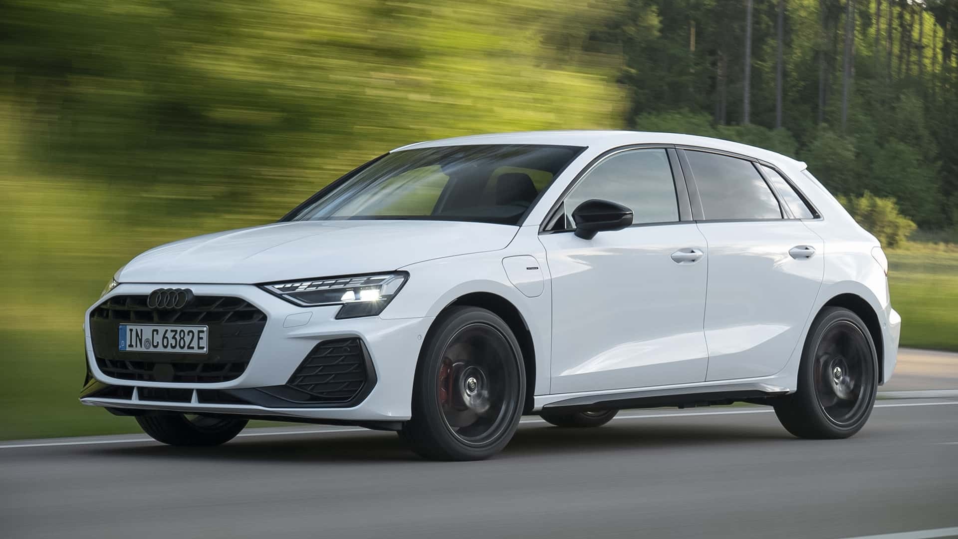 Carro Audi A3 branco em movimento, visto de lado, com faróis acesos, em uma estrada com árvores verdes borradas ao fundo
