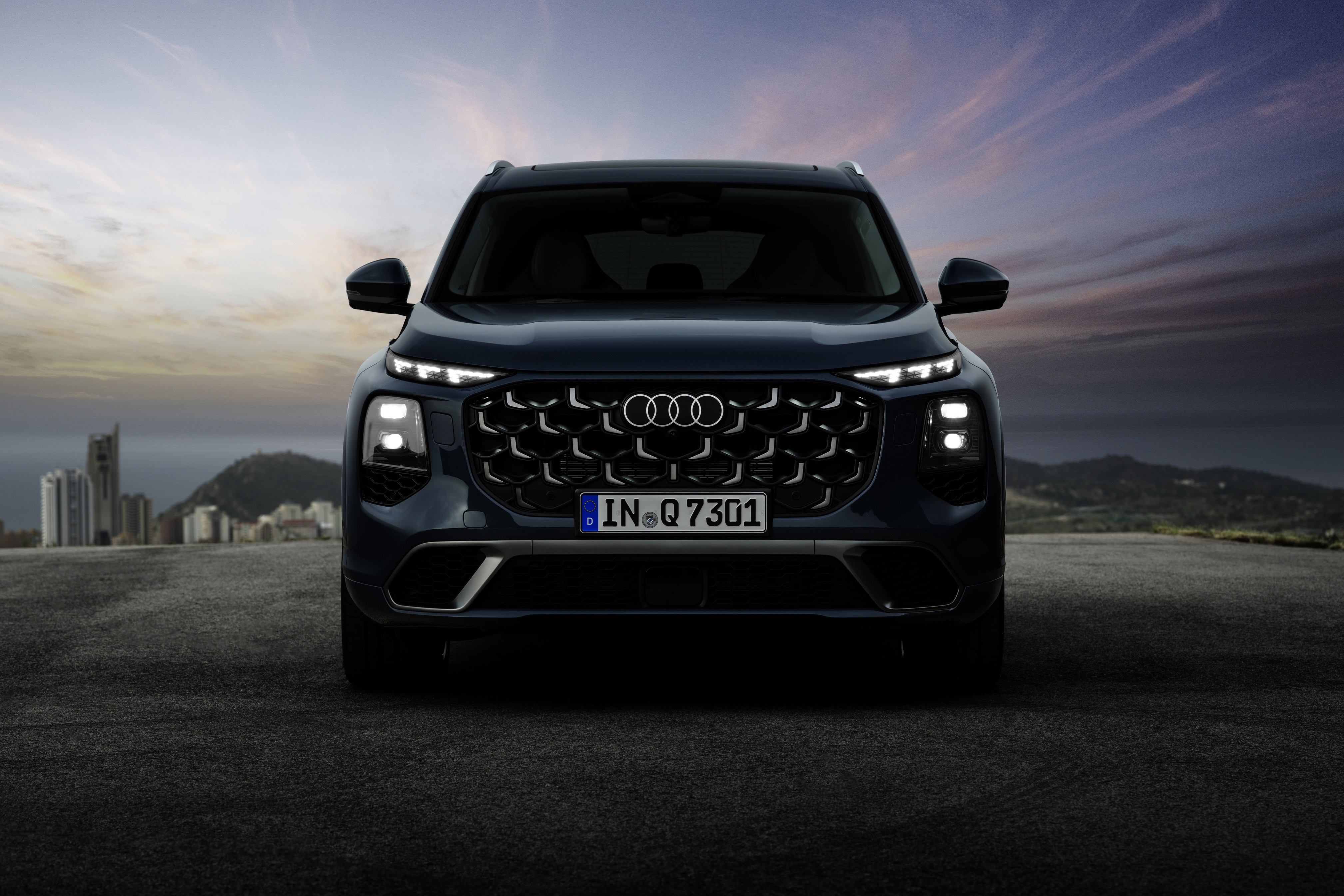 SUV Audi Q7 azul escuro com faróis e luzes diurnas acesas, grade frontal hexagonal e placa IN Q 7301, parado em asfalto escuro. Ao fundo, montanhas e prédios sob céu nublado ao entardecer