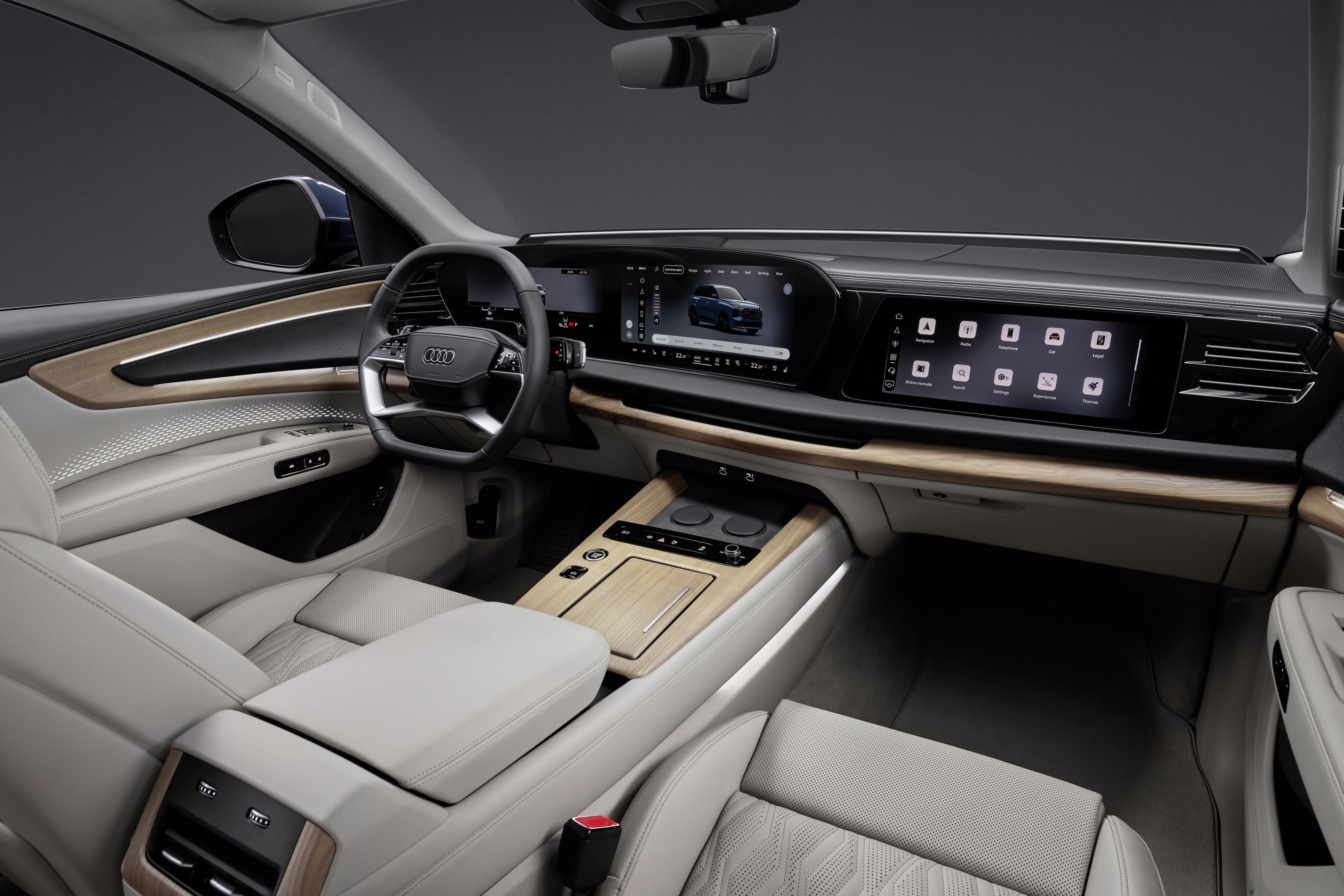 Interior de um carro Audi moderno, com bancos de couro cinza claro, painel em madeira clara e telas digitais. O volante tem o logotipo da Audi, e o console central possui botões e um porta-copos.