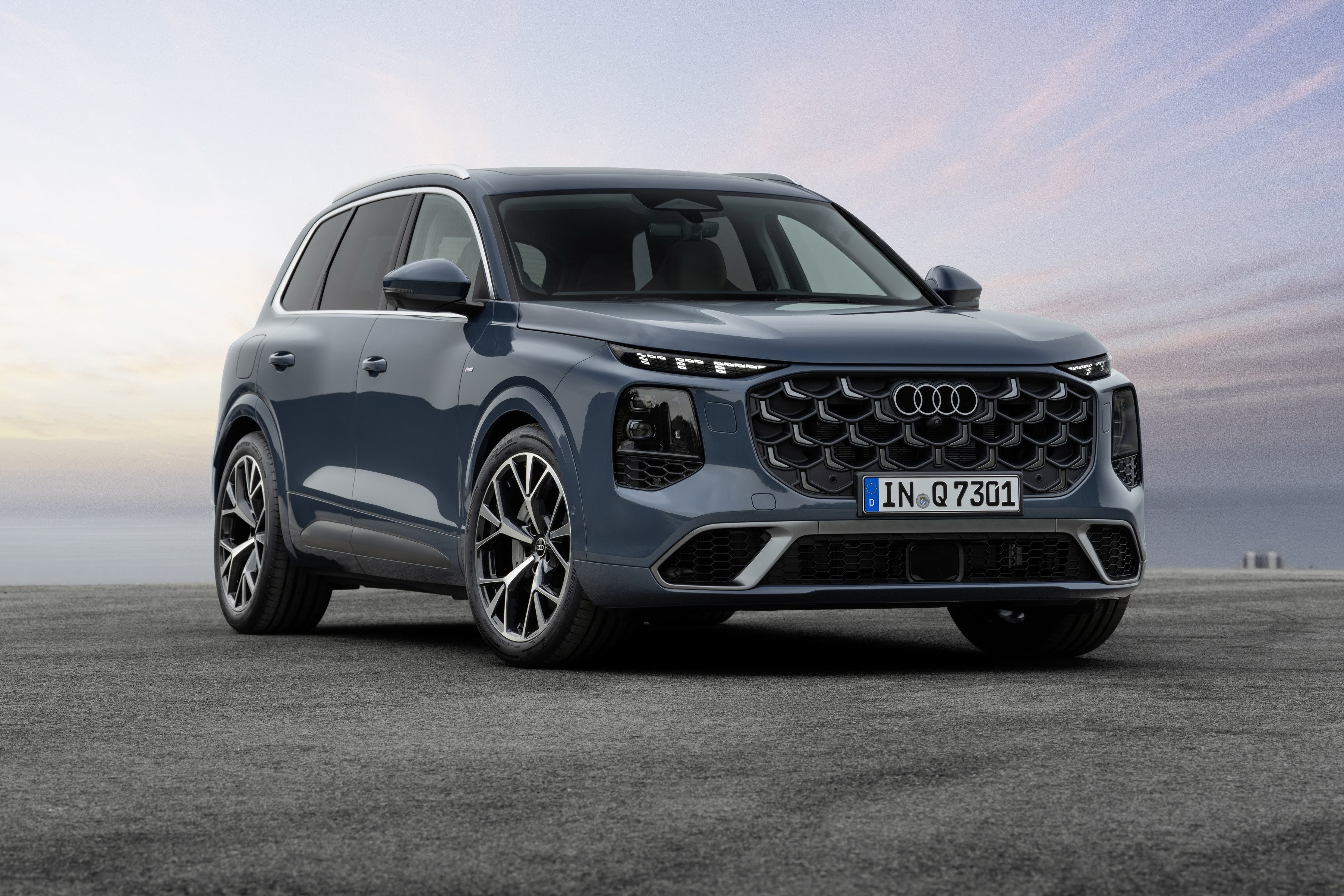 SUV Audi Q6 e-tron cinza-azulado, com grade frontal preta em padrão hexagonal e faróis LED divididos, estacionado em asfalto cinza sob céu claro com nuvens esparsas ao entardecer
