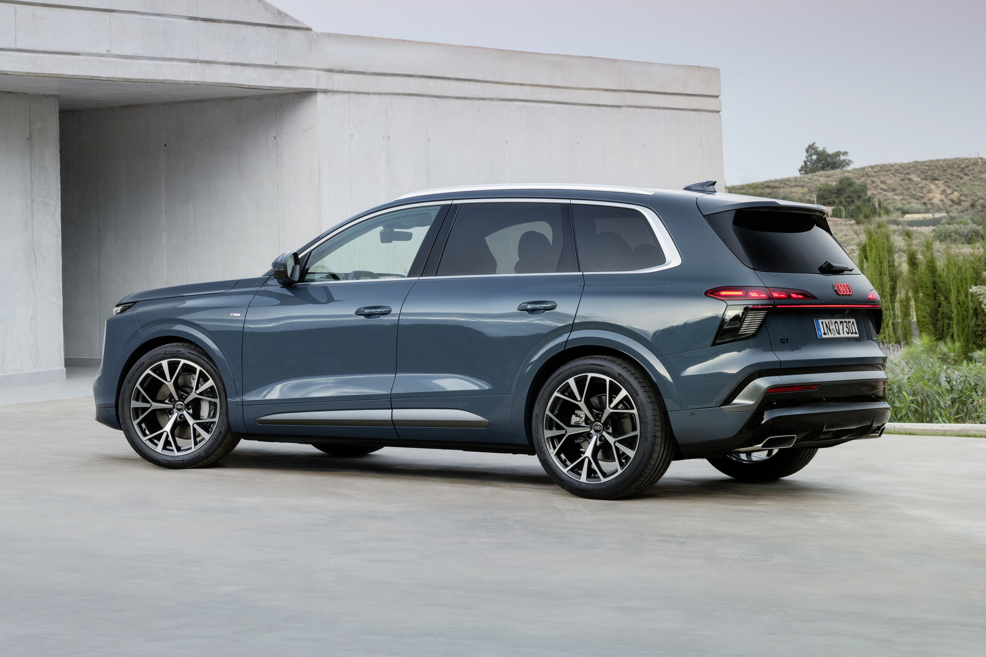 SUV Audi Q azul-escuro estacionado em uma superfície cinza, com um prédio branco moderno ao fundo e vegetação em um morro distante
