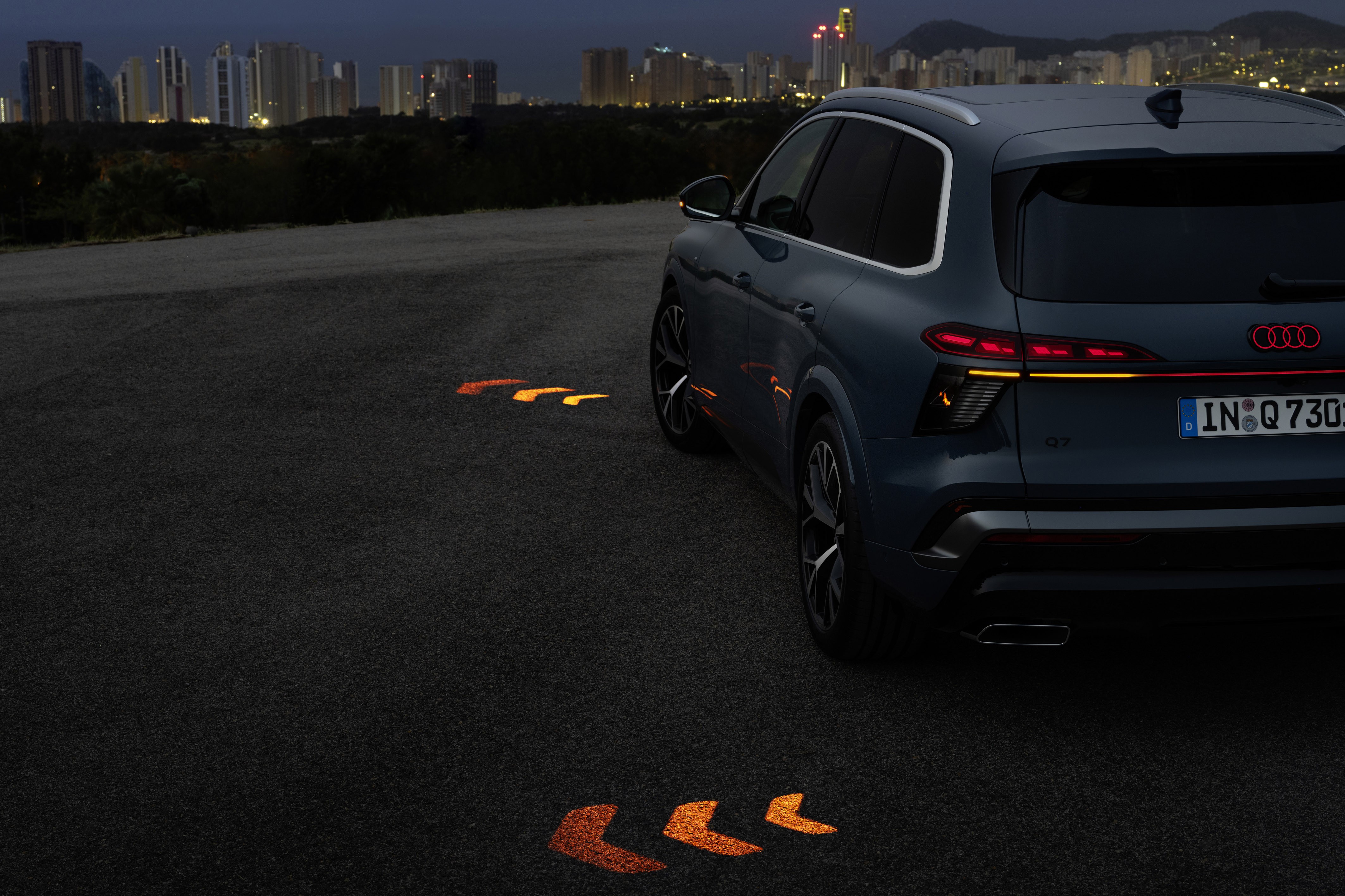 SUV Audi Q7 azul escuro estacionado em asfalto cinza escuro à noite, com setas luminosas laranja projetadas no chão. Ao fundo, uma cidade iluminada com prédios altos