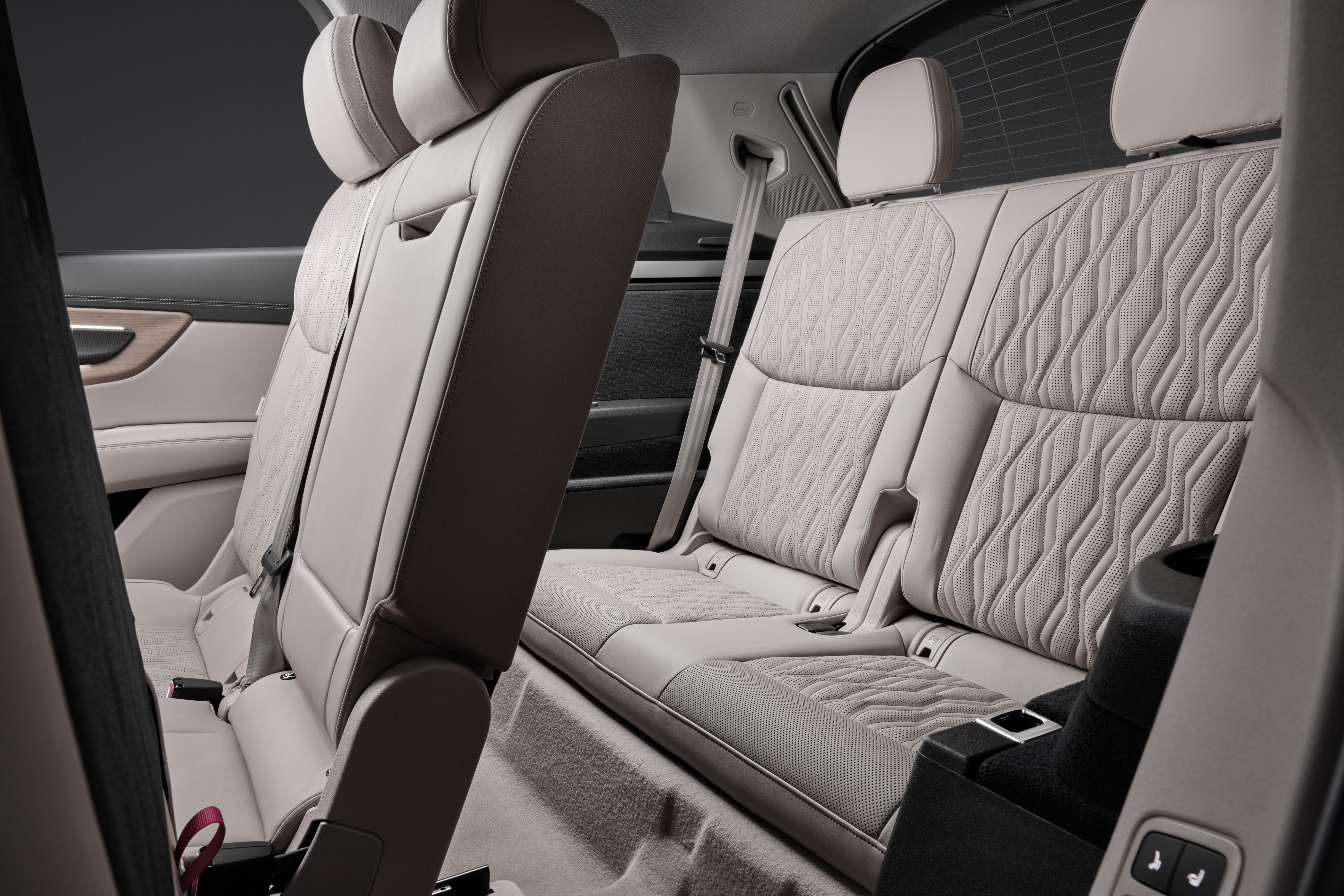 Interior de carro com bancos de couro bege claro, estofamento com padrão geométrico em relevo. O banco traseiro direito está rebatido, mostrando o espaço para bagagem. Cintos de segurança visíveis, e um porta-copos preto no console lateral. Detalhes em madeira na porta.