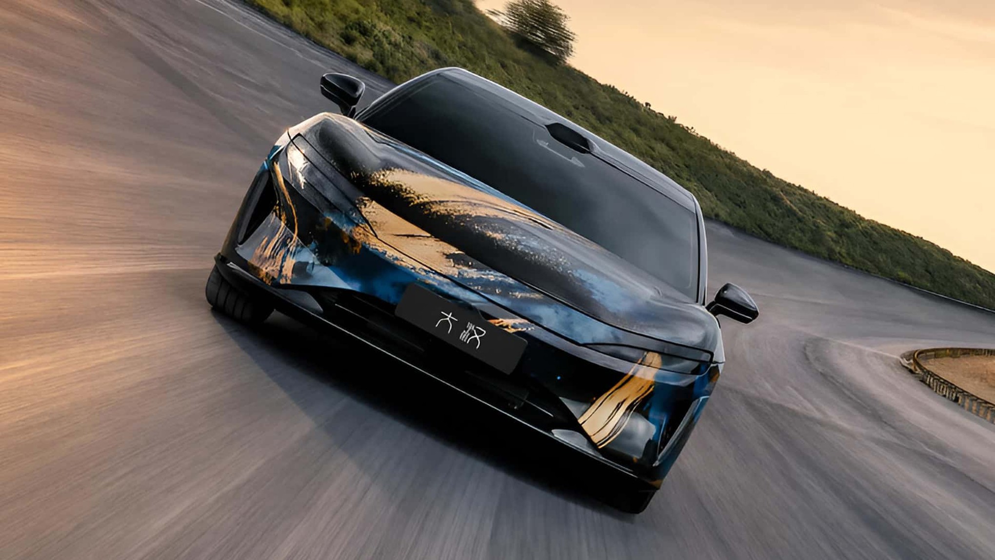 Carro esportivo preto com detalhes em azul e dourado, como pinceladas artísticas, em uma estrada sinuosa com colina verde ao fundo, sob céu claro e alaranjado