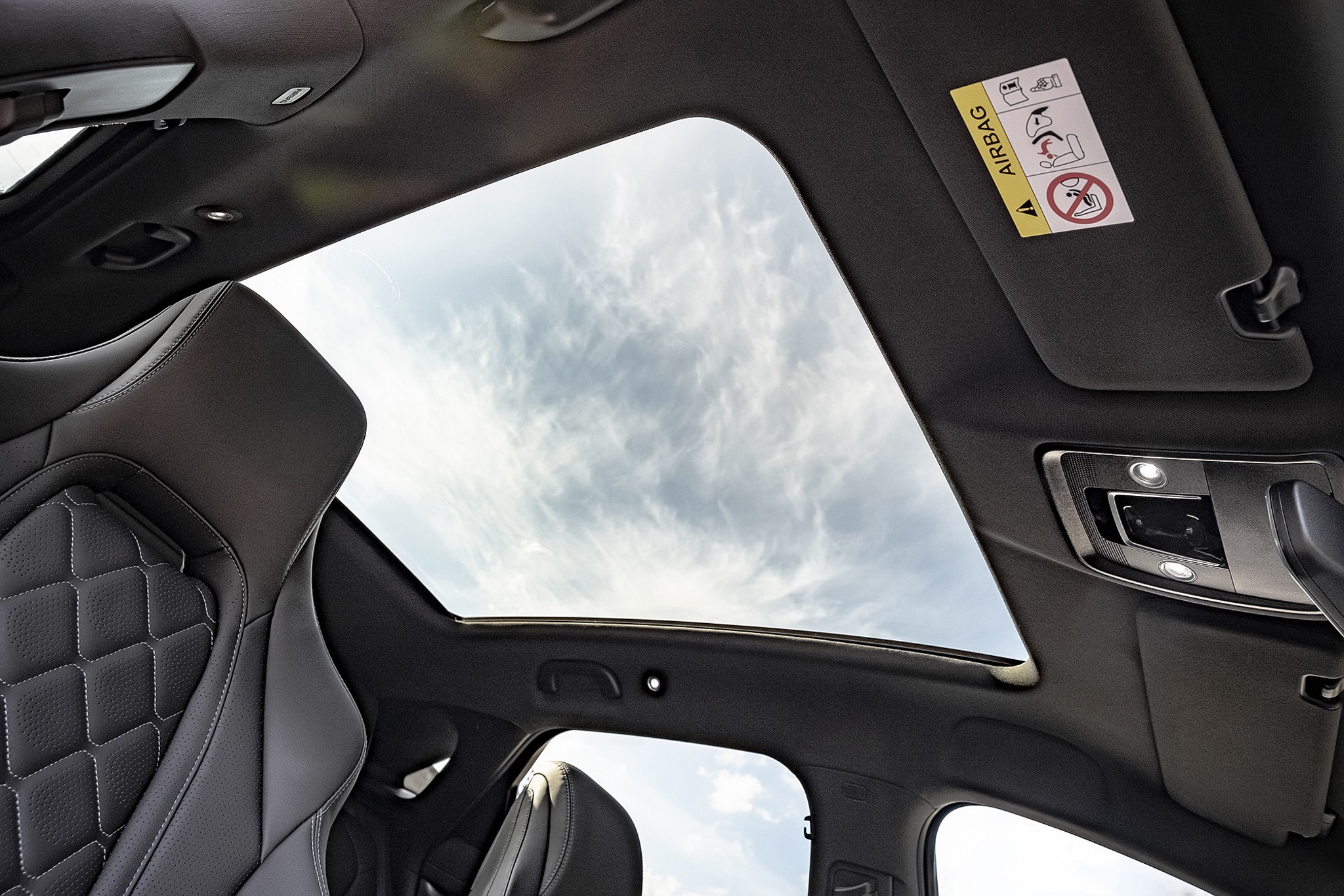 Interior de um carro com teto solar panorâmico, mostrando o céu azul com nuvens esparsas. À esquerda, parte do banco de couro preto com costura em losangos. À direita, o console de luzes internas e o para-sol com adesivo de aviso do airbag