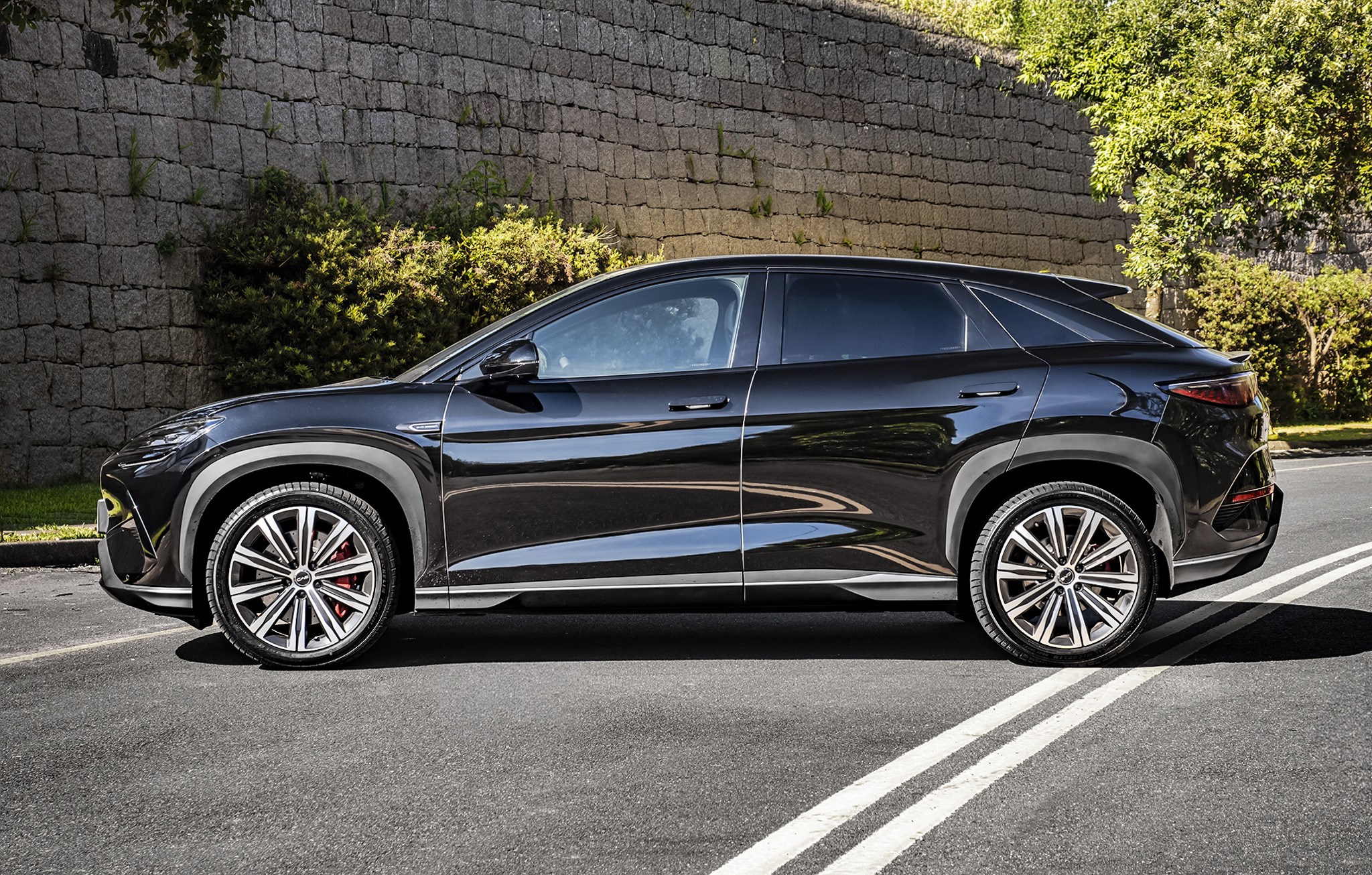 SUV preto brilhante estacionado de perfil em uma rua asfaltada, com uma parede de pedra e vegetação ao fundo. O carro tem rodas esportivas prateadas com detalhes vermelhos nos freios e acabamentos cinzas nas caixas de roda. Duas faixas brancas paralelas marcam o asfalto em primeiro plano