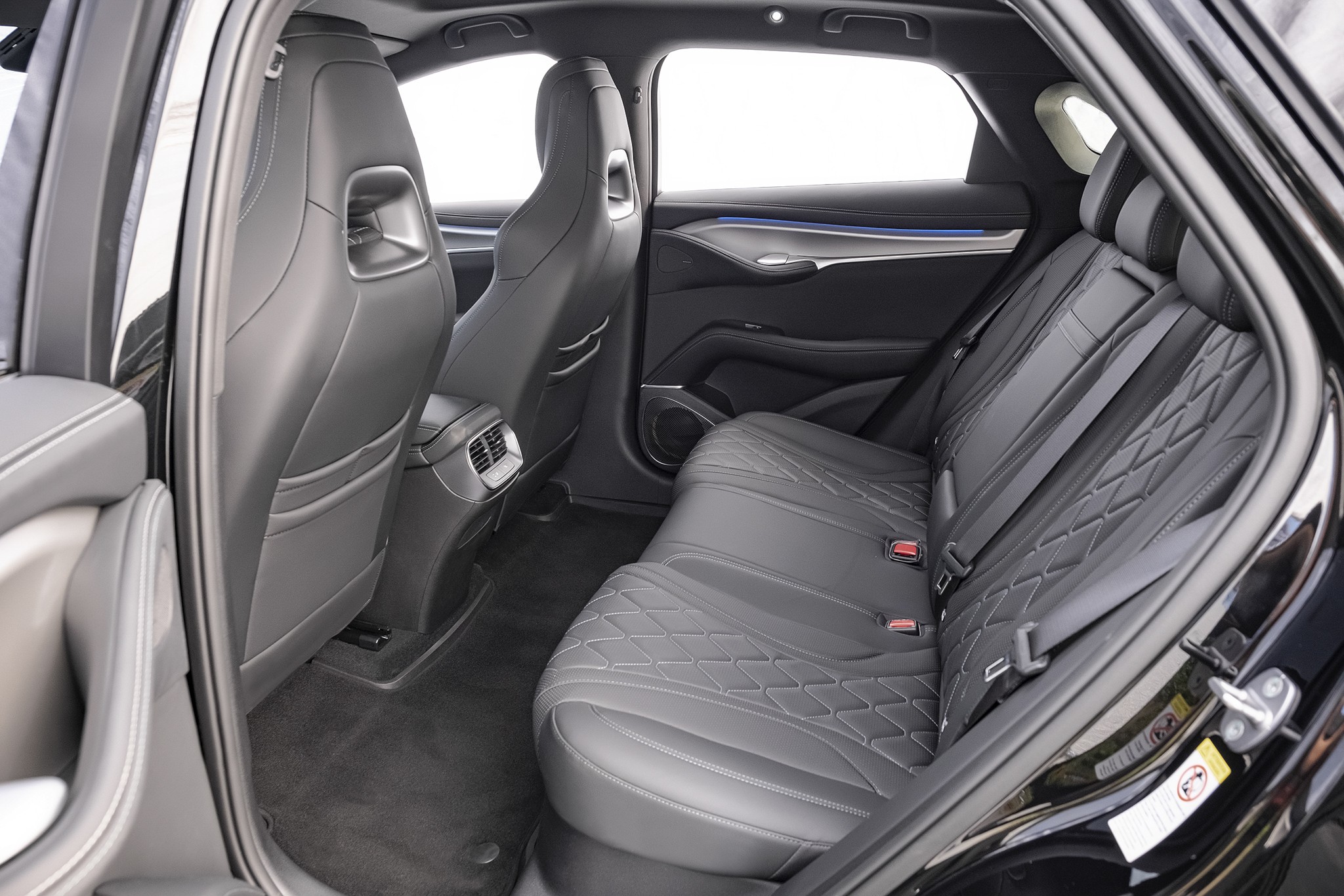 Interior de um carro com bancos traseiros de couro preto e costura em losango, vistos da porta traseira aberta. Os bancos dianteiros também são pretos, com detalhes prateados nos encostos. O piso é escuro e as janelas mostram um fundo branco