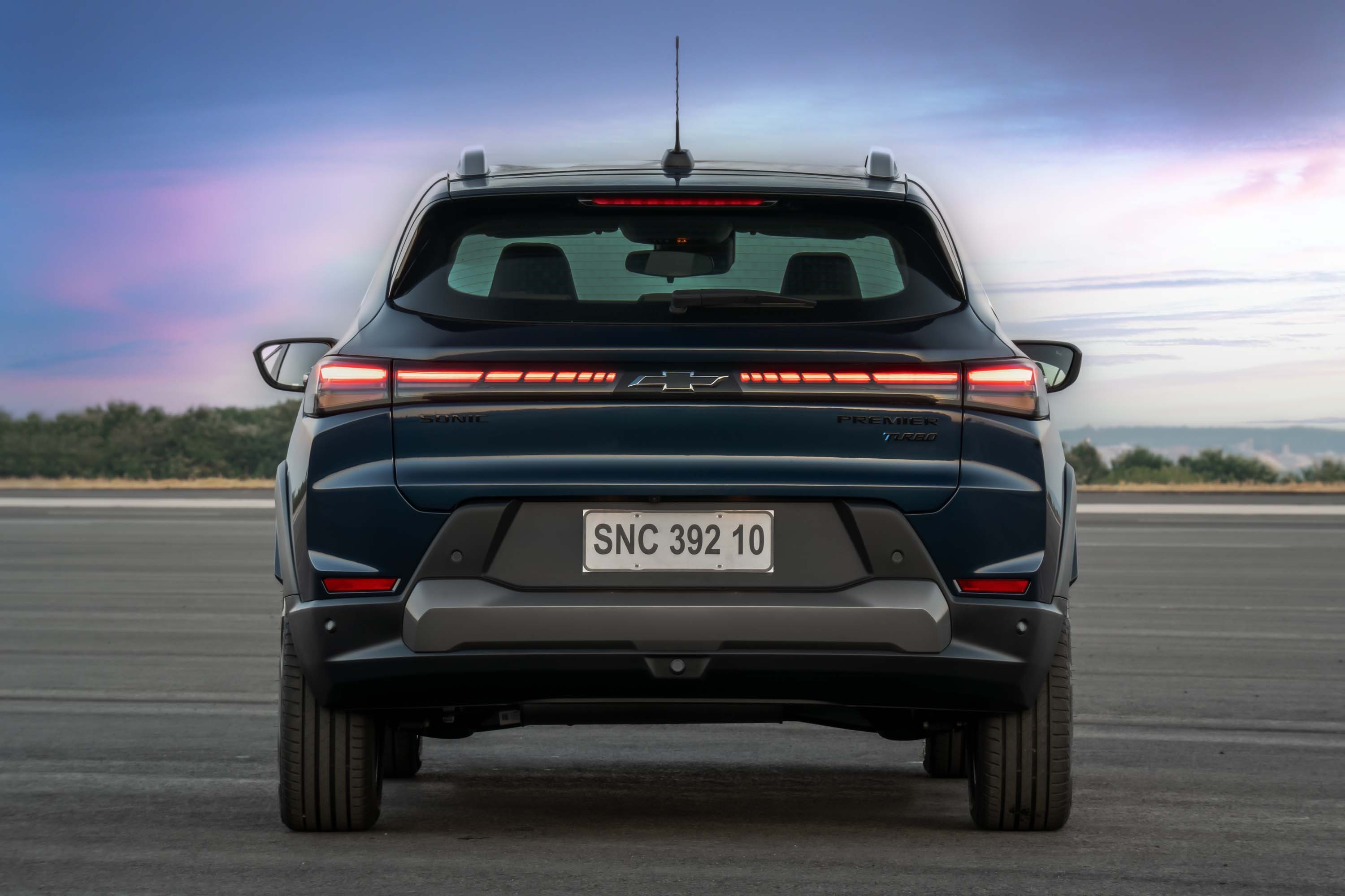 Traseira de um carro SUV azul-escuro, com lanternas horizontais finas e acesas, sobre as quais está o emblema Chevrolet. A placa branca exibe SNC 392 10. O para-choque traseiro é cinza-escuro, e o veículo está em uma pista de asfalto, com um céu azul e nuvens ao fundo