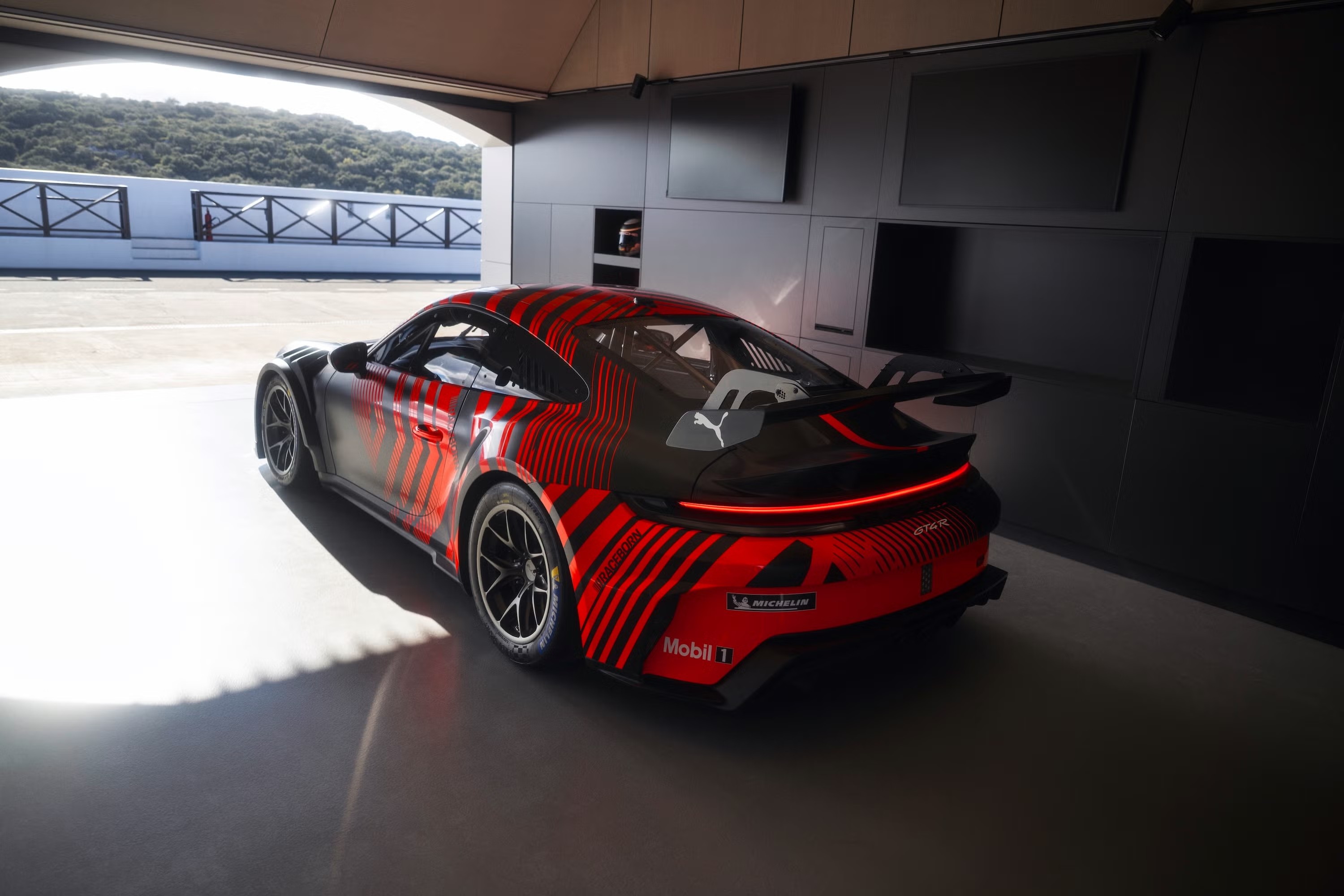 Carro esportivo preto e vermelho com listras diagonais, visto de trás, estacionado em uma garagem escura com a porta aberta para uma pista de corrida e montanhas ao fundo