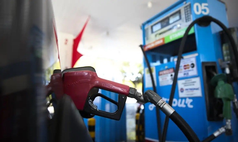 Bico de combustível vermelho inserido no tanque de um veículo, com uma bomba de gasolina azul e branca ao fundo, em um posto de combustível