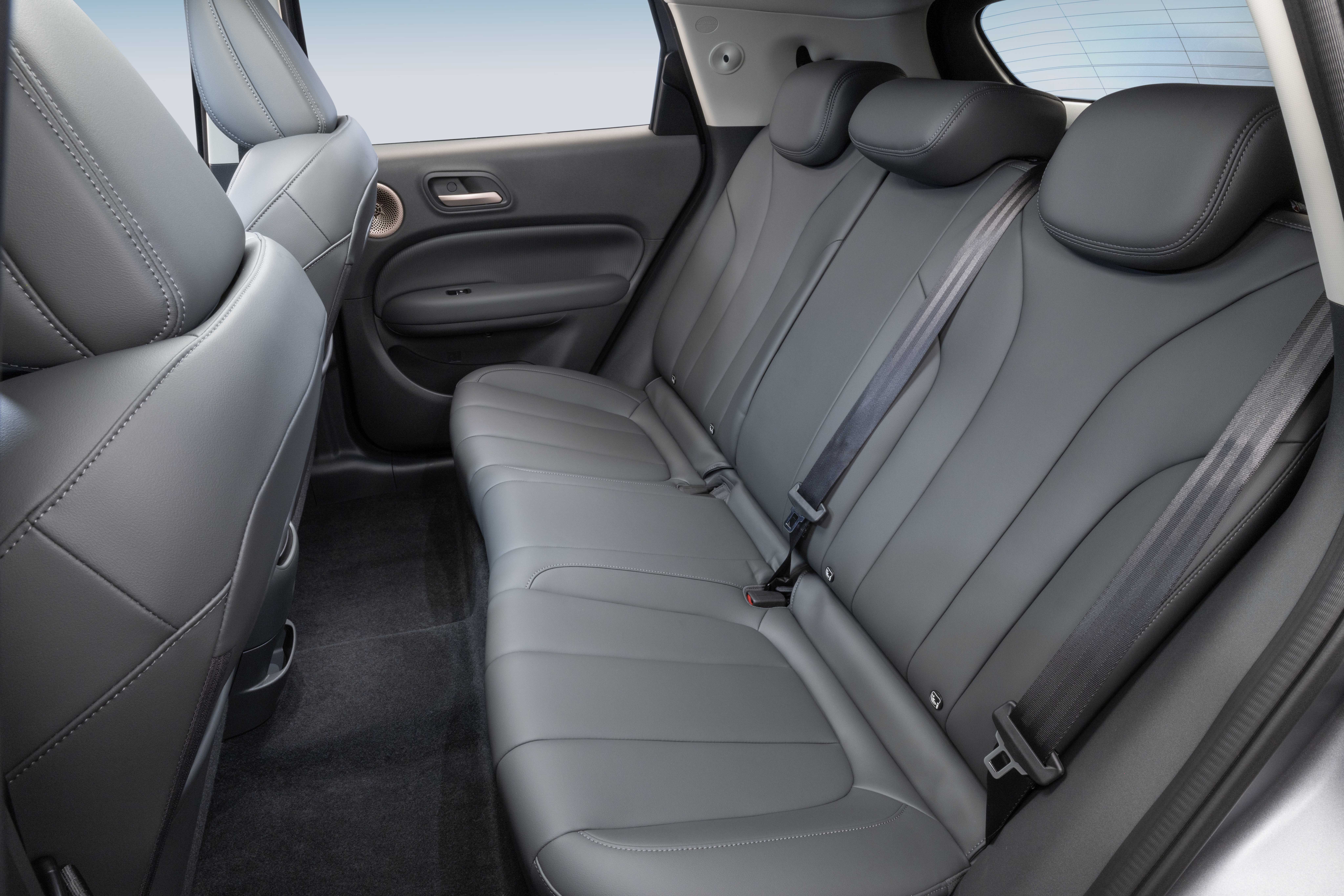 Interior de carro com bancos traseiros de couro cinza escuro, com encostos de cabeça e cintos de segurança visíveis. O banco do passageiro dianteiro, também de couro cinza, aparece parcialmente à esquerda. O carpete é preto e a porta lateral tem detalhes escuros e maçaneta cromada
