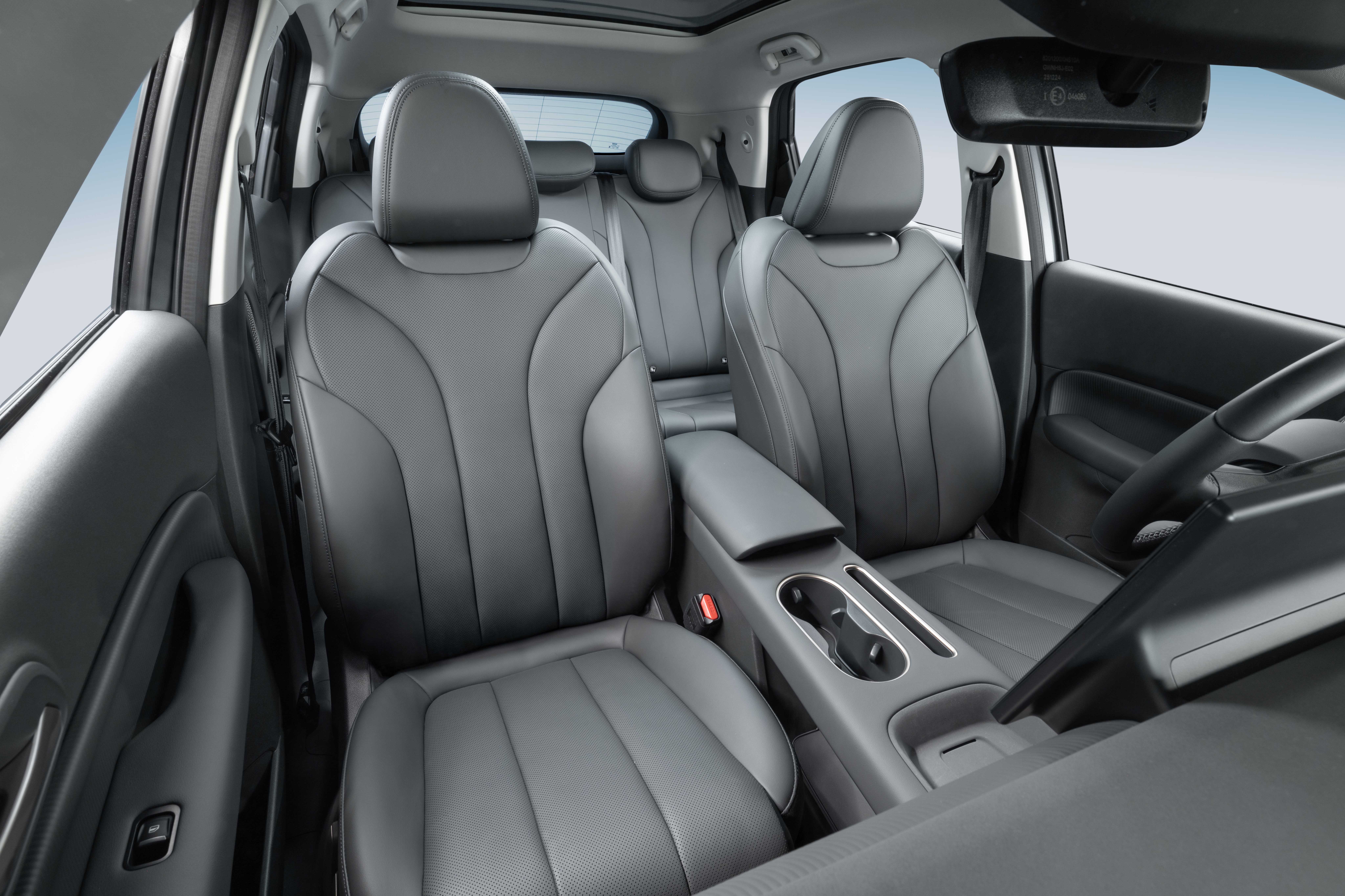 Interior de um carro com bancos de couro cinza escuro, incluindo os assentos dianteiros e parte dos traseiros. O console central possui porta-copos e apoio de braço. O volante e o retrovisor interno são visíveis, e o teto solar está aberto, mostrando o céu azul