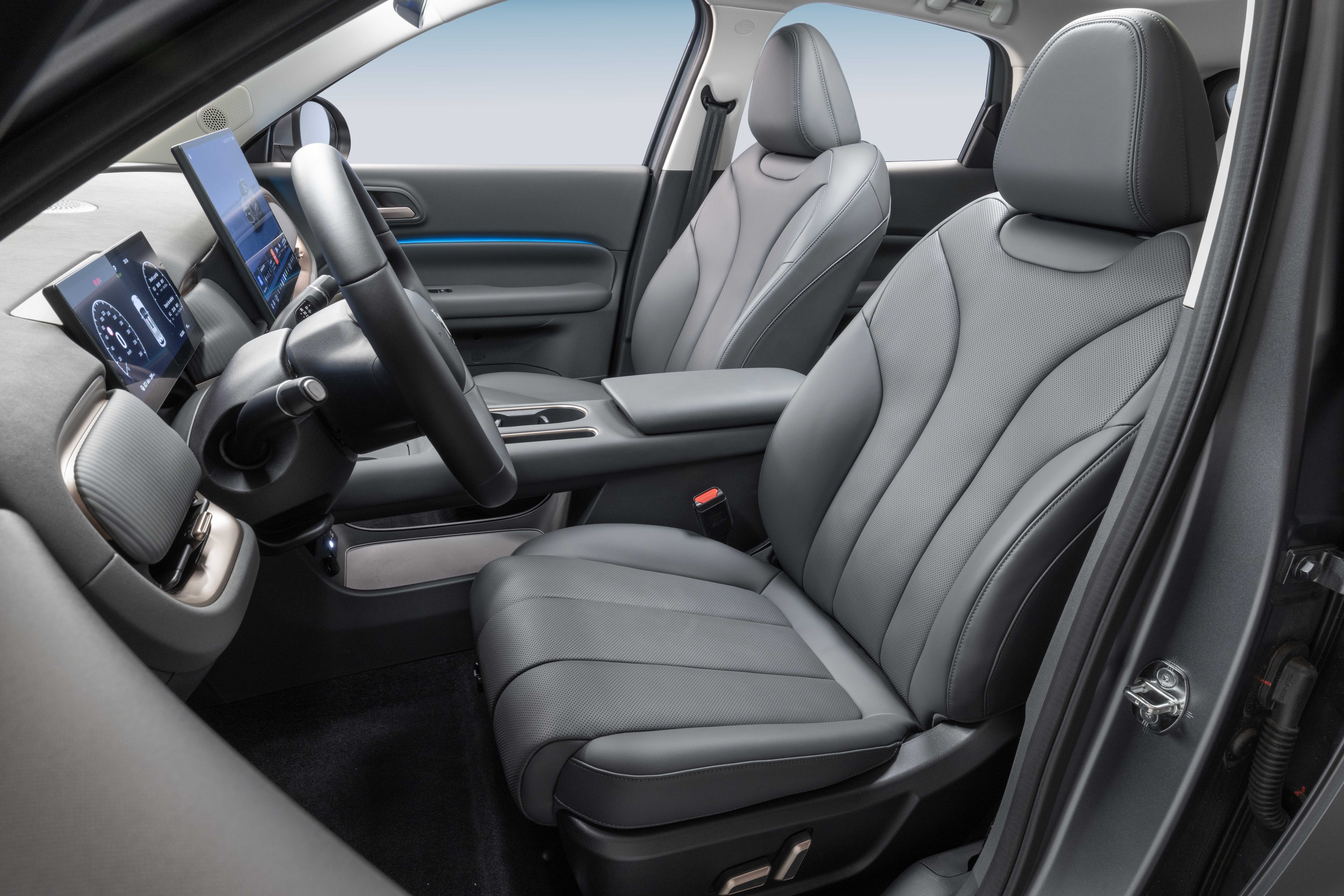 Interior de carro moderno com bancos de couro cinza escuro, volante à esquerda e duas telas digitais no painel, uma para o motorista e outra central. Detalhe azul claro na porta.