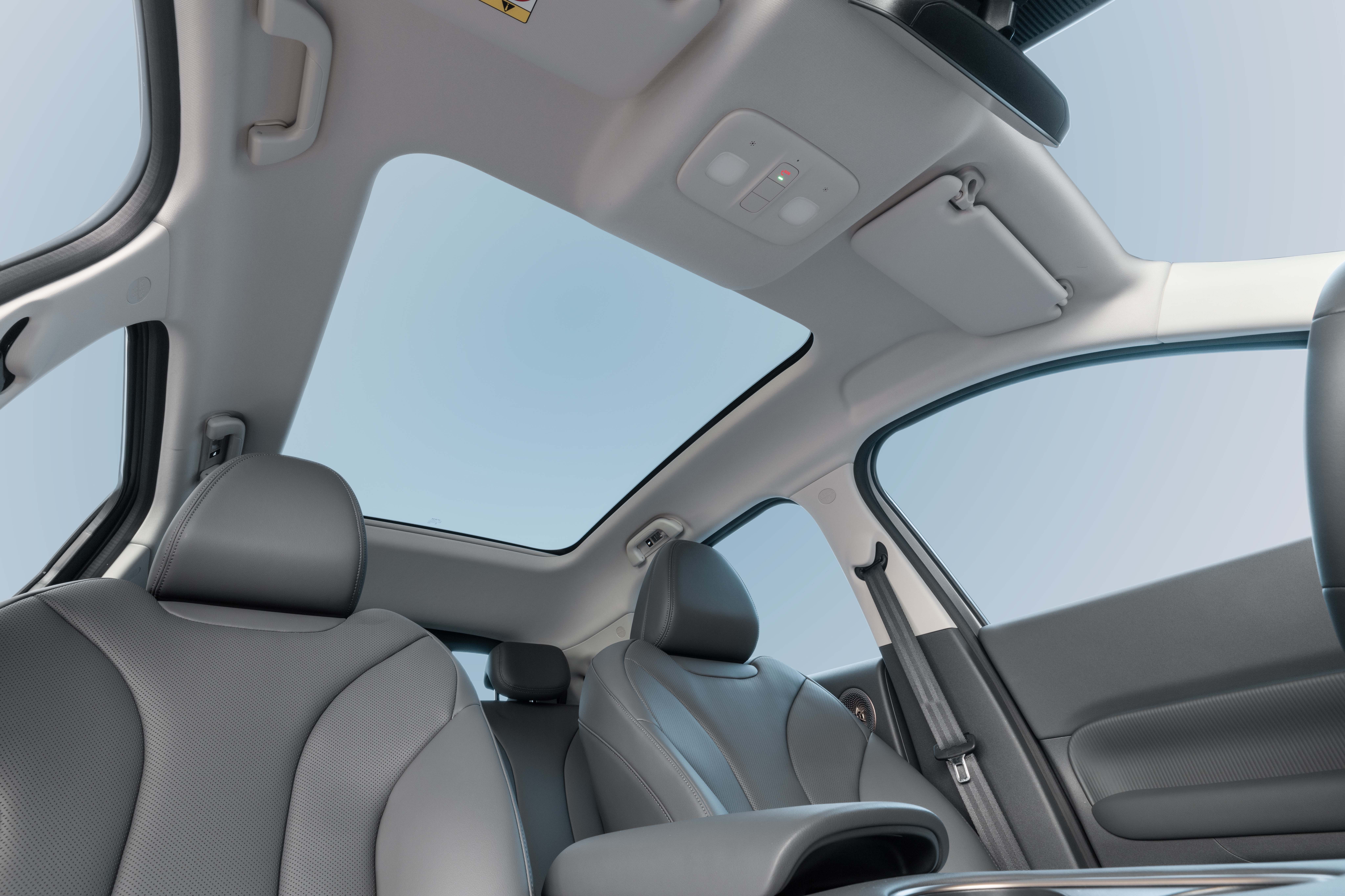 Interior de um carro com teto solar panorâmico, mostrando o céu azul. Os bancos dianteiros e traseiros são de couro cinza, com detalhes perfurados. O console central e as laterais das portas também são visíveis, com um cinto de segurança preto na lateral direita