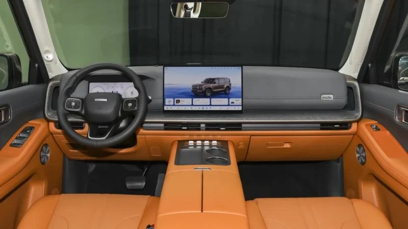 Interior de um carro moderno com estofamento e detalhes em couro marrom-claro. O painel cinza tem uma tela multimídia grande exibindo um SUV cinza e um volante multifuncional preto. O console central possui porta-copos e botões de controle.