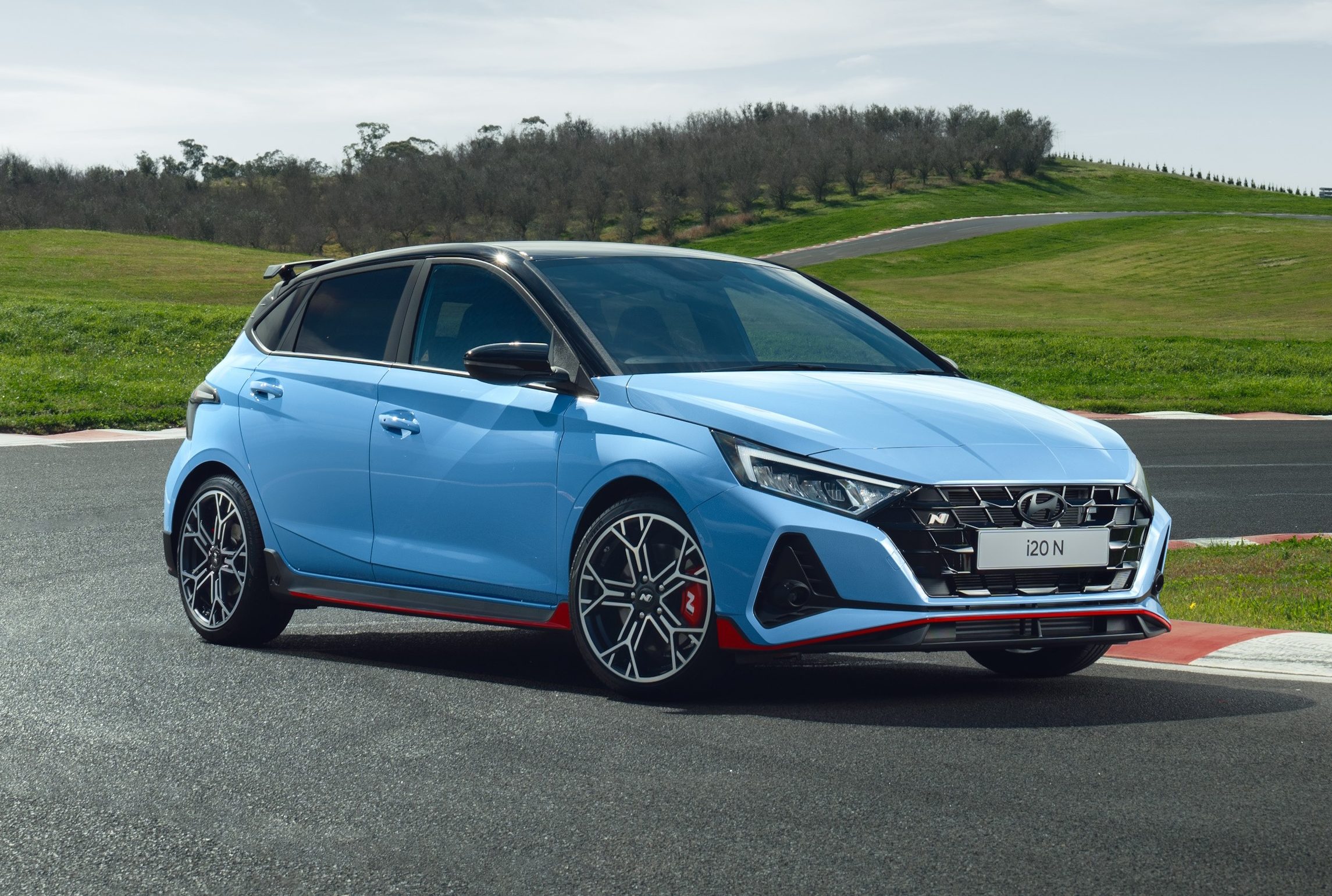 Hatchback azul claro com teto preto e detalhes vermelhos, parado em uma pista de corrida com grama verde e céu azul ao fundo