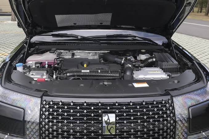 Motor de um carro preto com o capô aberto, revelando componentes como o reservatório de fluido rosa, bateria e mangueiras. A grade frontal possui um padrão de losangos e um emblema central prateado. O veículo está estacionado em um piso de blocos cinzas, com uma estrada e árvores ao fundo