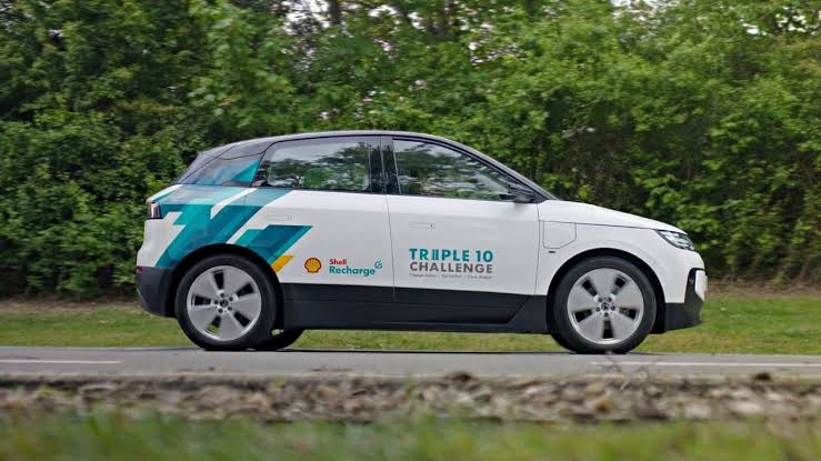 Carro elétrico branco com detalhes azuis e verdes, logotipos Shell Recharge e Triple 10 Challenge, parado em uma estrada asfaltada com vegetação ao fundo