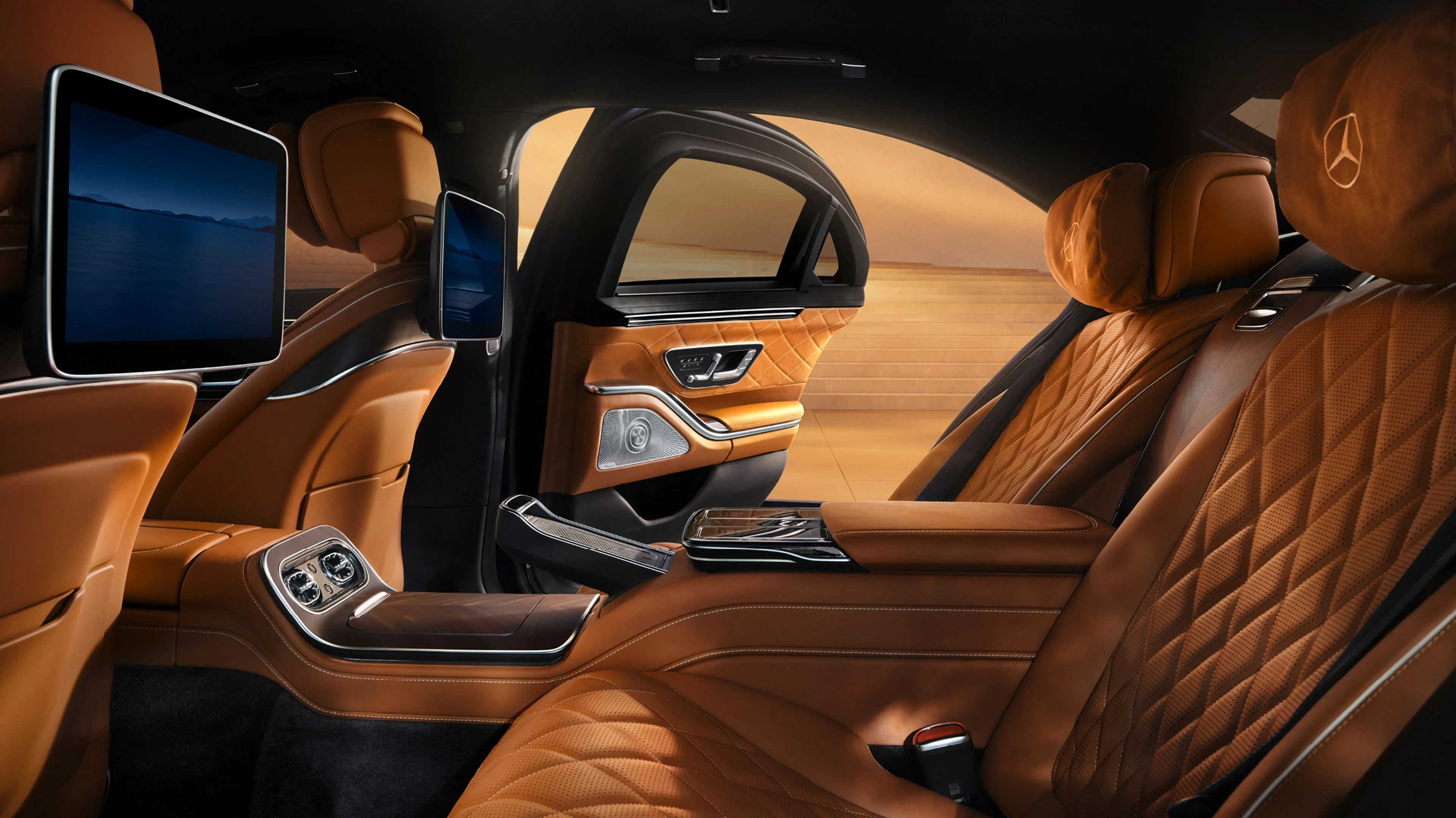 Interior luxuoso de um carro com bancos de couro marrom acolchoado, telas multimídia nos encostos e detalhes metálicos. A porta traseira está aberta, revelando um cenário desfocado de deserto ao fundo
